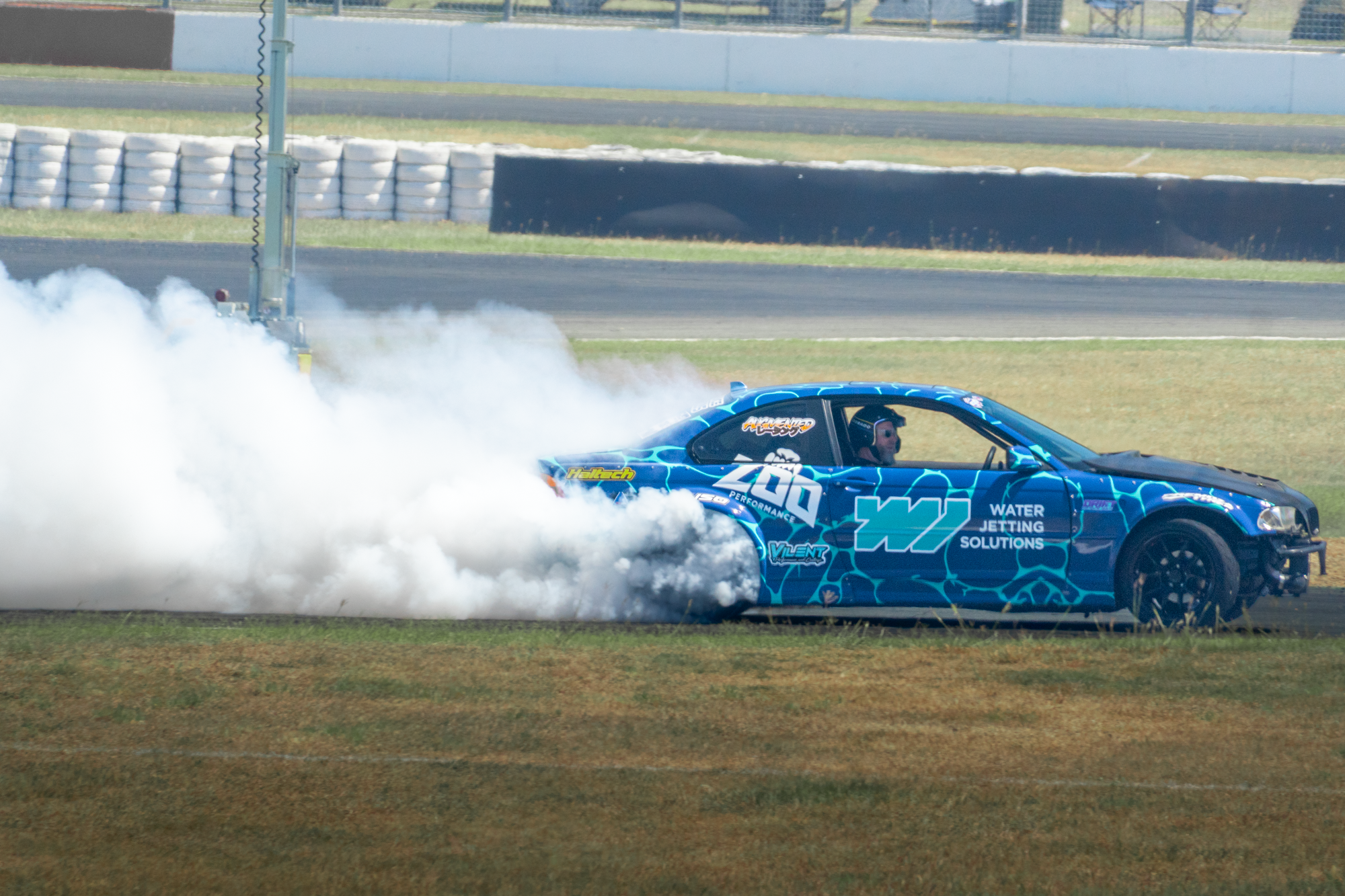 A blue race car with the number 40 is making a burnout on a race track, producing a large cloud of white smoke. The driver is wearing sunglasses, and there are tire barriers and a fence in the background.