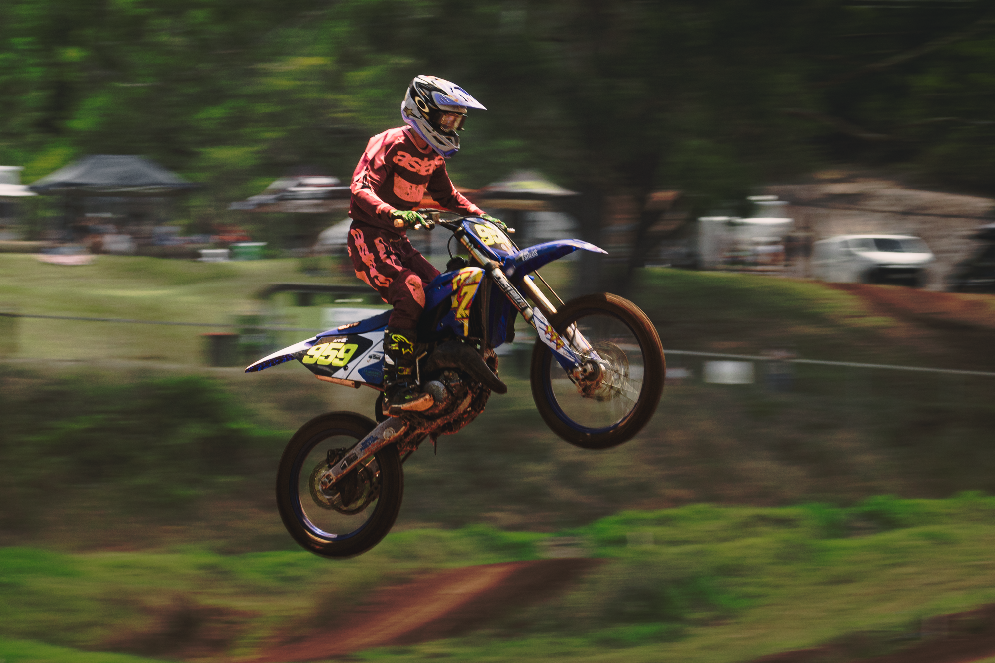 A motocross rider in red gear and a helmet riding a blue dirt bike through a dirt track outdoors during daytime.