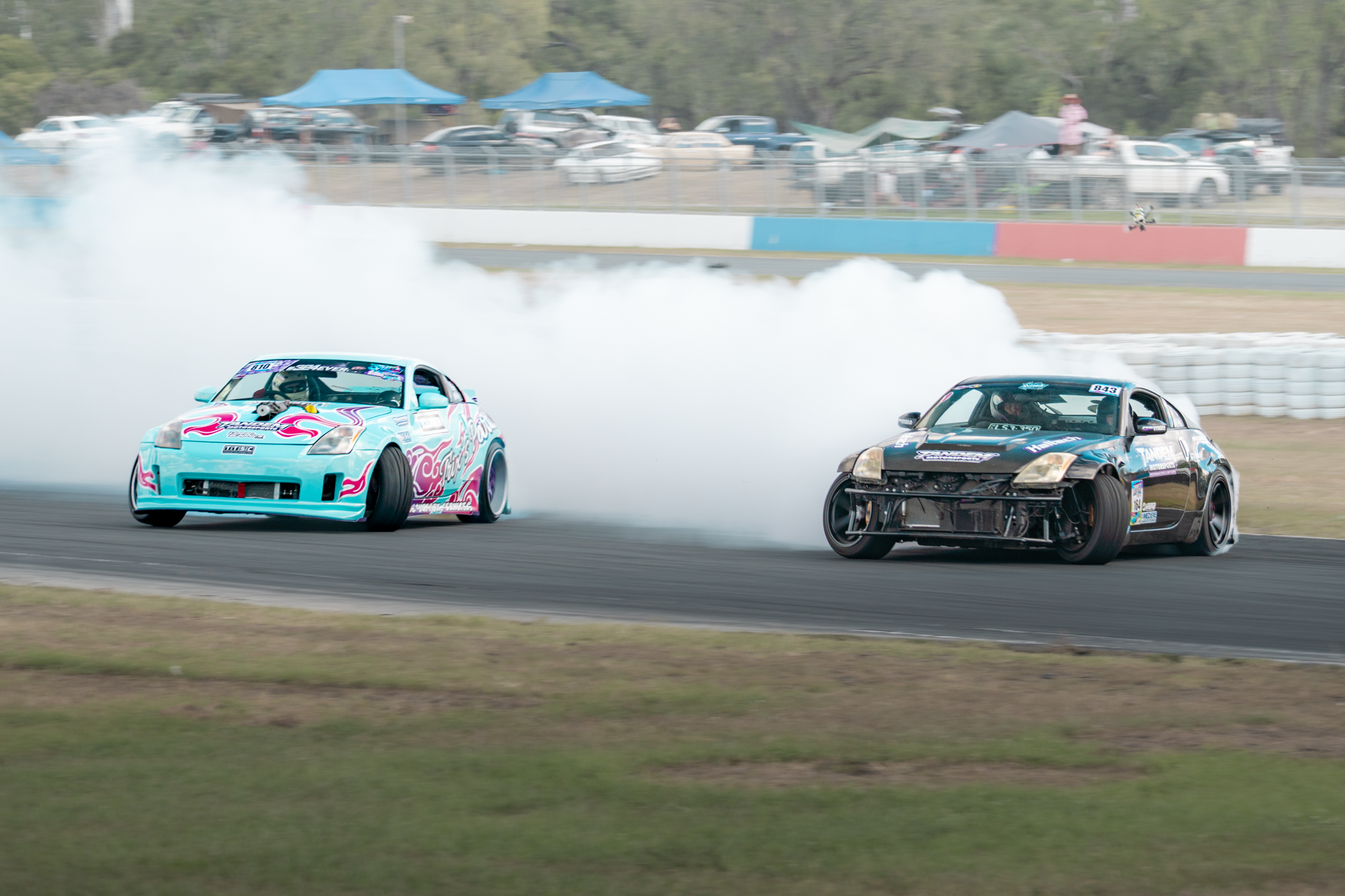 Two race cars drifting on a race track, creating smoke behind them, with cars and spectators visible in the background.