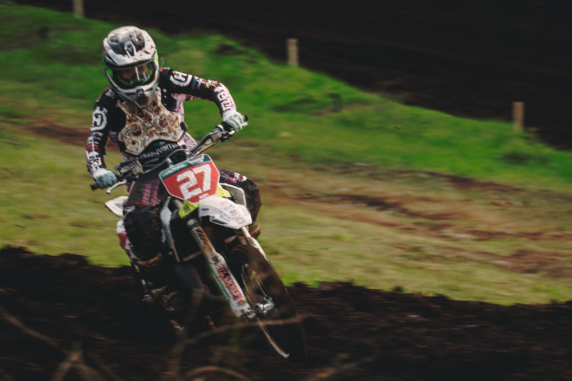 A motocross rider wearing a helmet and protective gear is riding a dirt bike on a dirt track surrounded by grass, with motion blur indicating high speed.