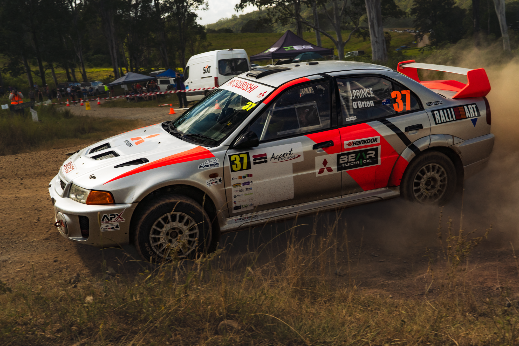A rally car with the number 37 on the side is driving on a dirt road, kicking up dust, during a race event. In the background, there is a crowd of people, tents, and vehicles among trees.