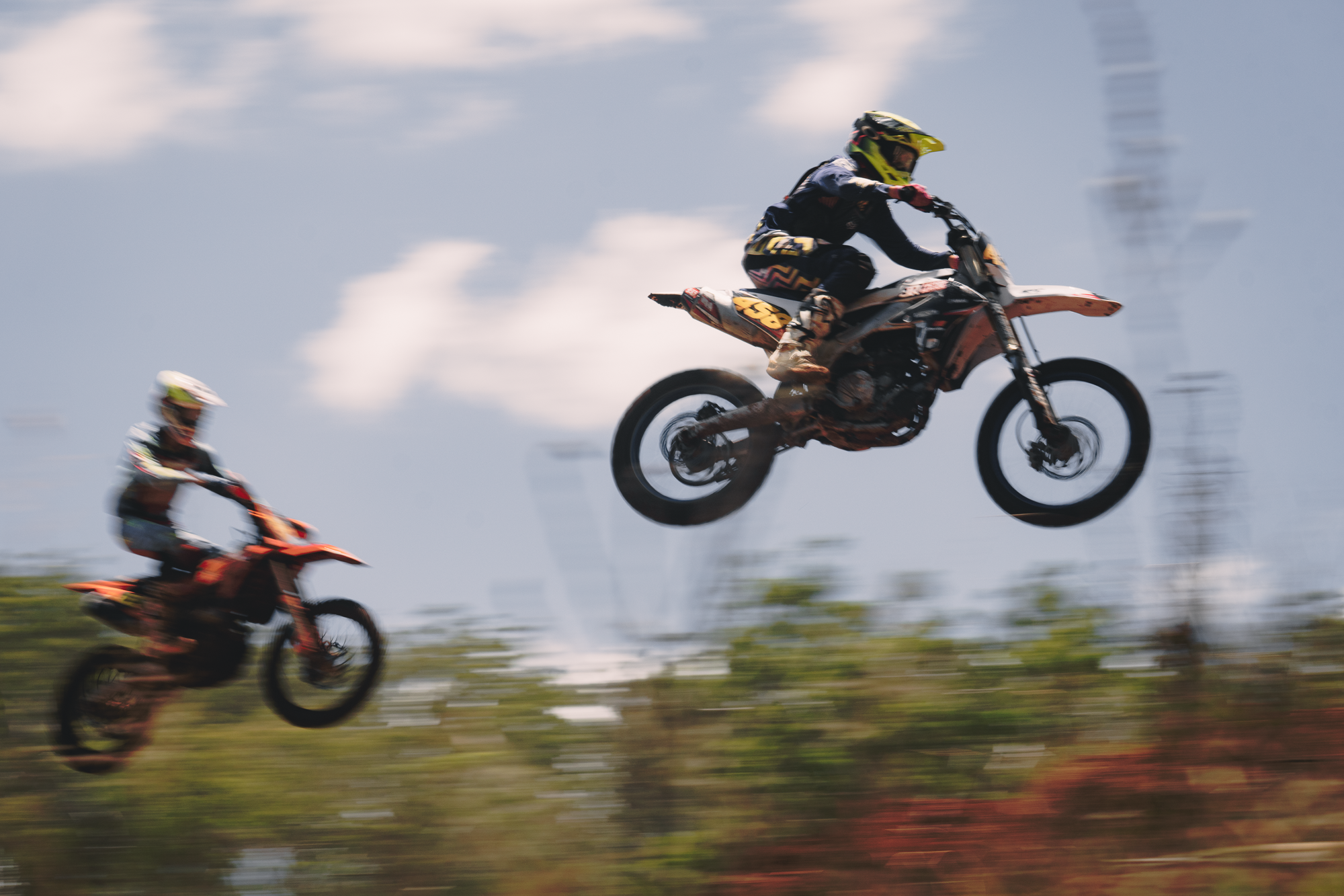 Two motocross riders in protective gear racing on a dirt track, with one rider airborne and the other riding on the ground, with a cloudy sky in the background.