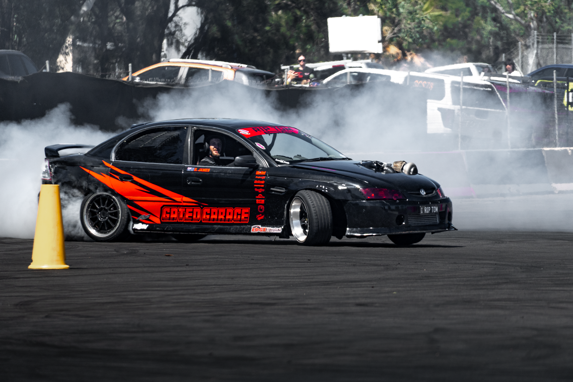 A black sports car with orange and red graphics, labeled 'GATED GARAGE,' is drifting on a race track, generating smoke from the tires. The driver, wearing a helmet, is smiling and looking out the window. Orange cone in the foreground and a background