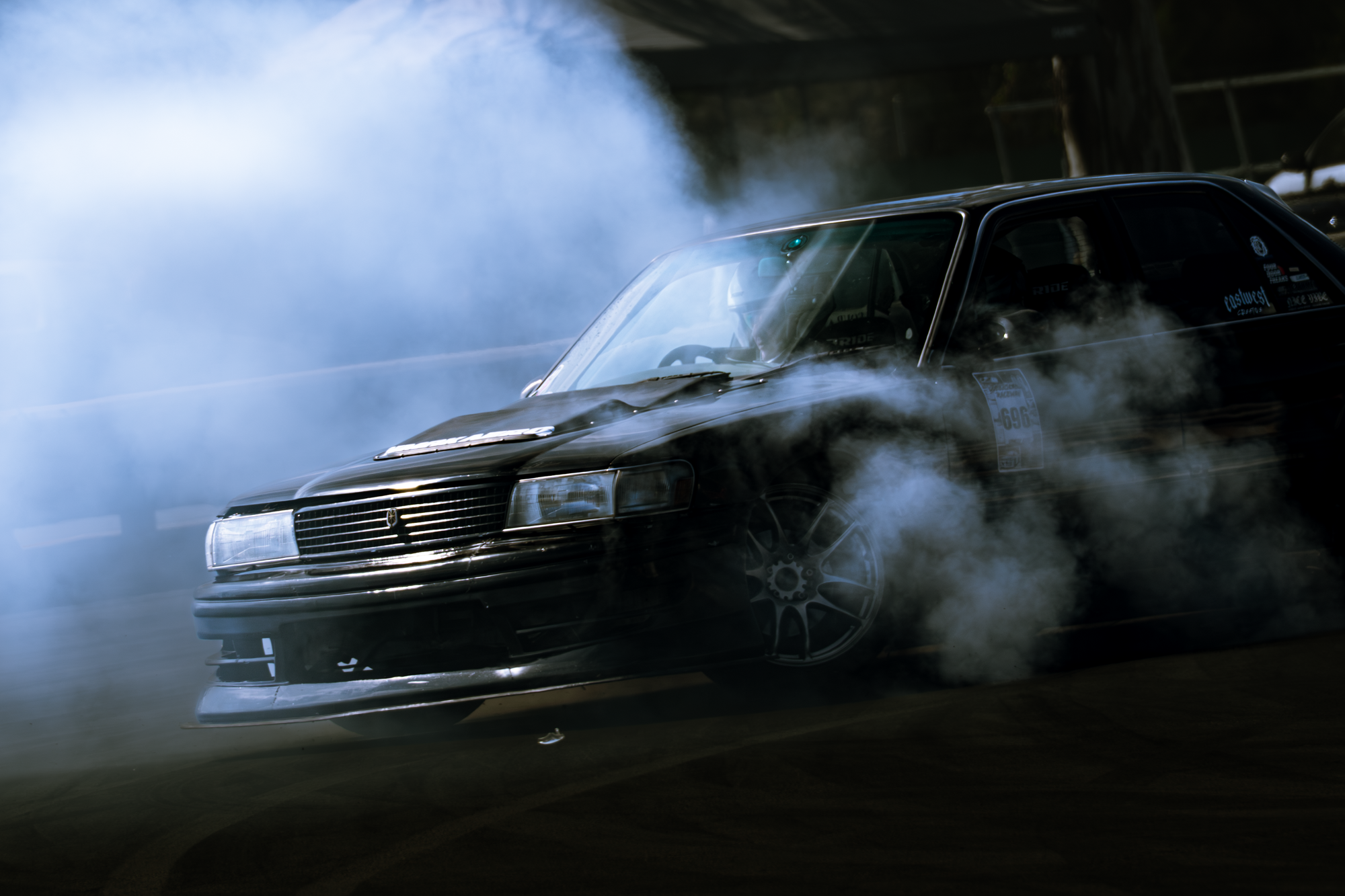 Black car performing a burnout surrounded by smoke or tire smoke on a dark racing or drifting track.