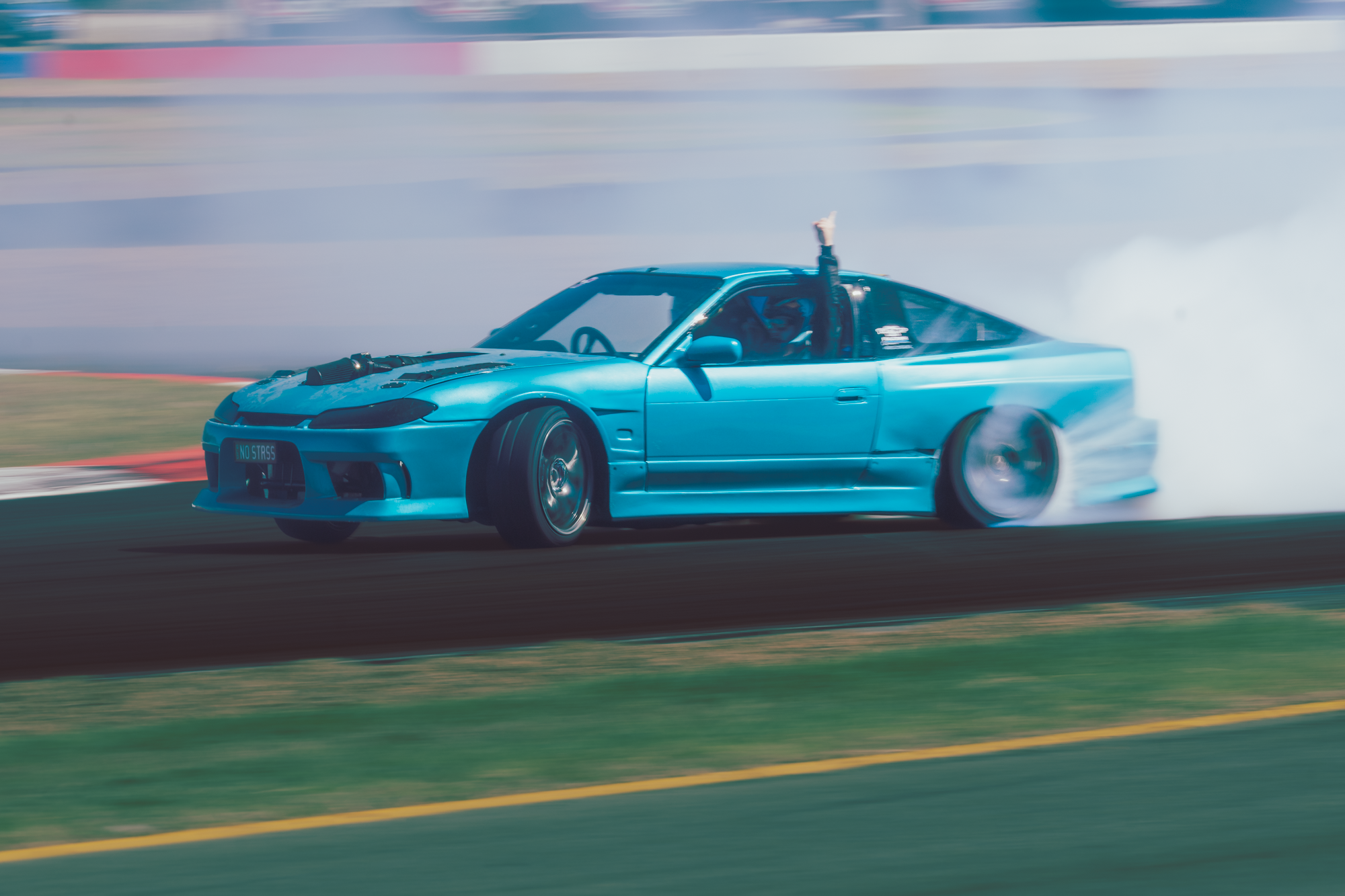 A blue sports car driving on a race track with tire smoke and a person raising their arm through the window.