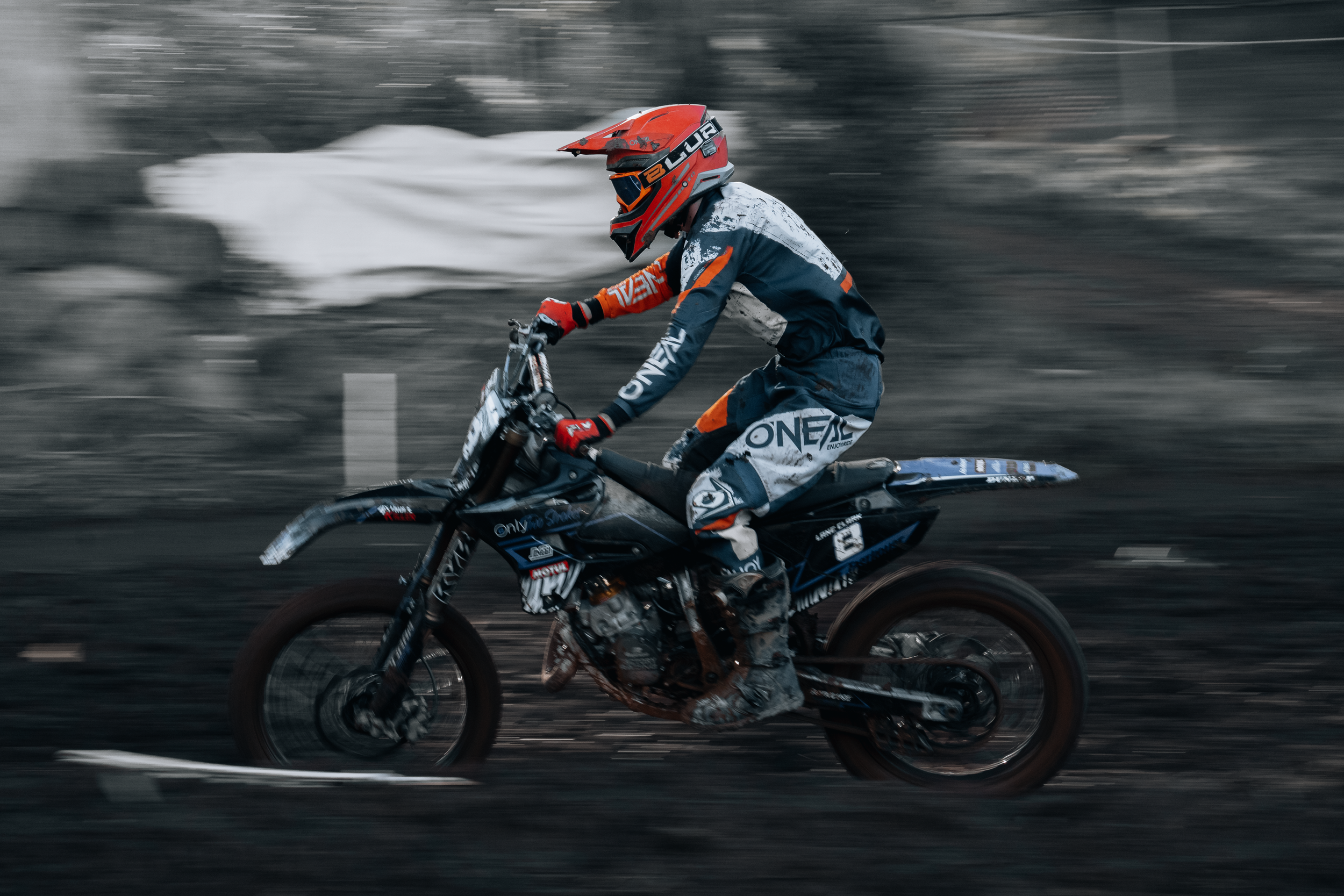 A person dressed in motocross gear riding a dirt bike at high speed on a dirt trail, with motion blur in the background.