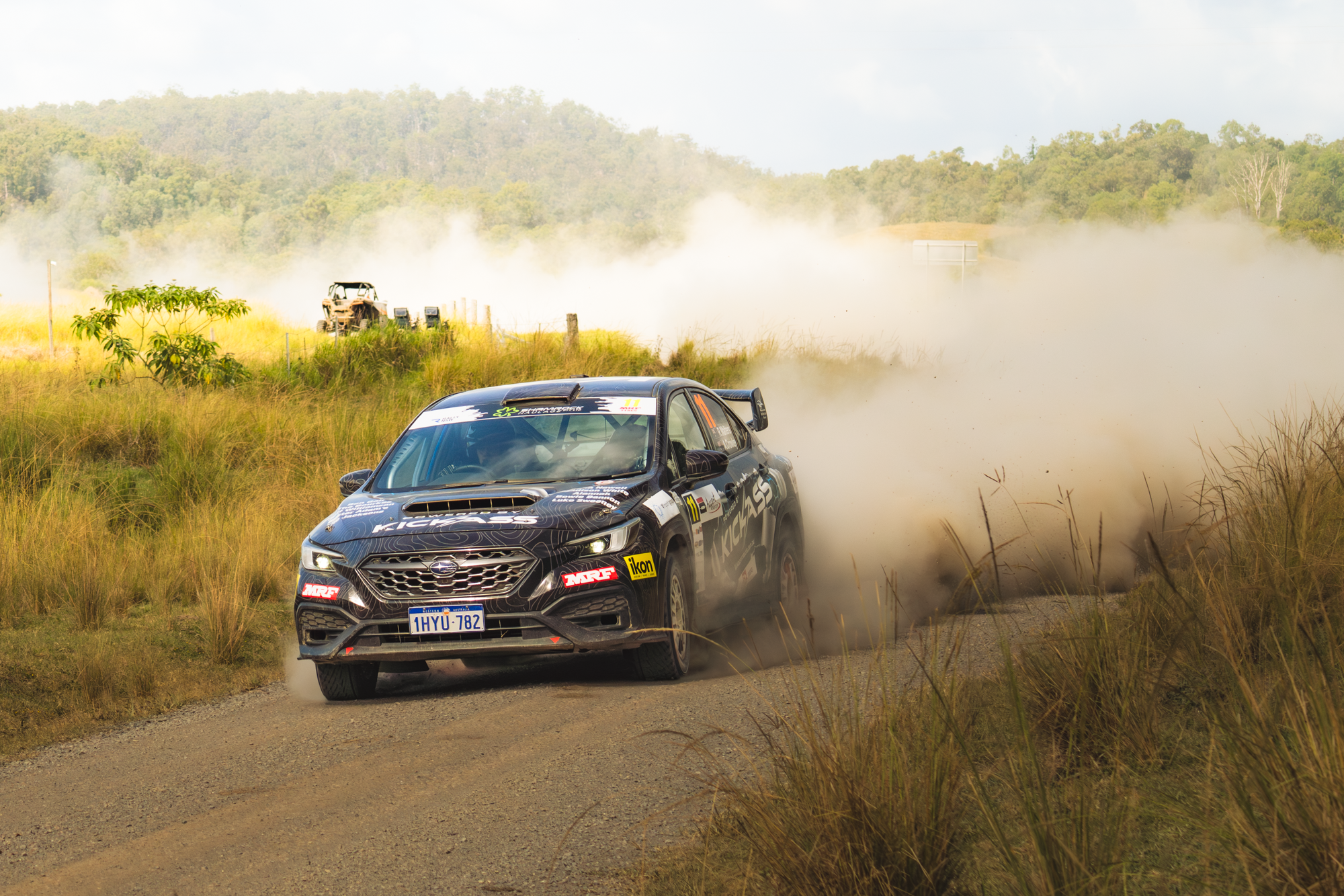 A black rally car driving on an unpaved road, kicking up dust with a rural landscape in the background, including trees and grass.