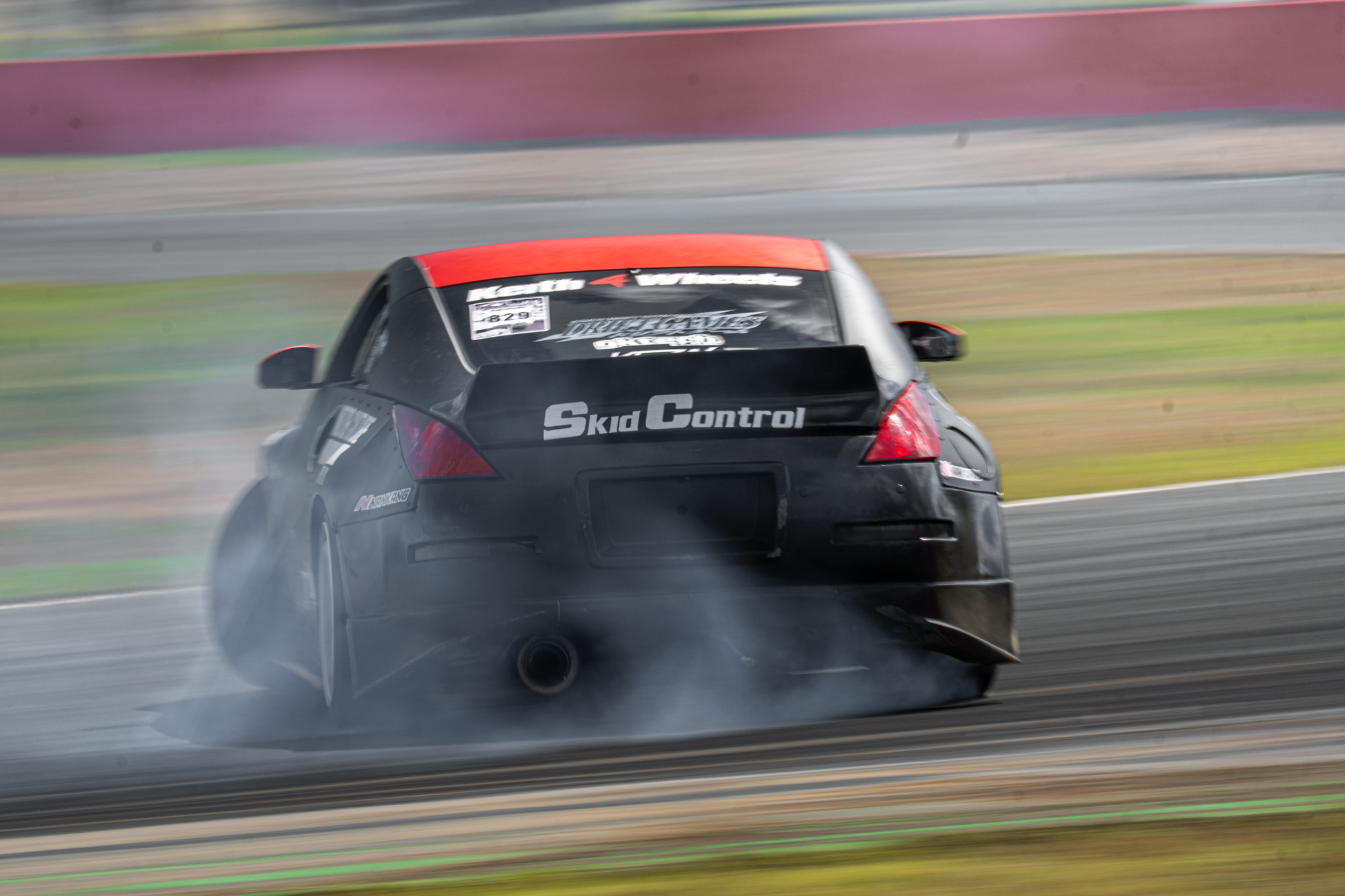 A black car with red accents racing on a track, creating tire smoke with a blurred background.