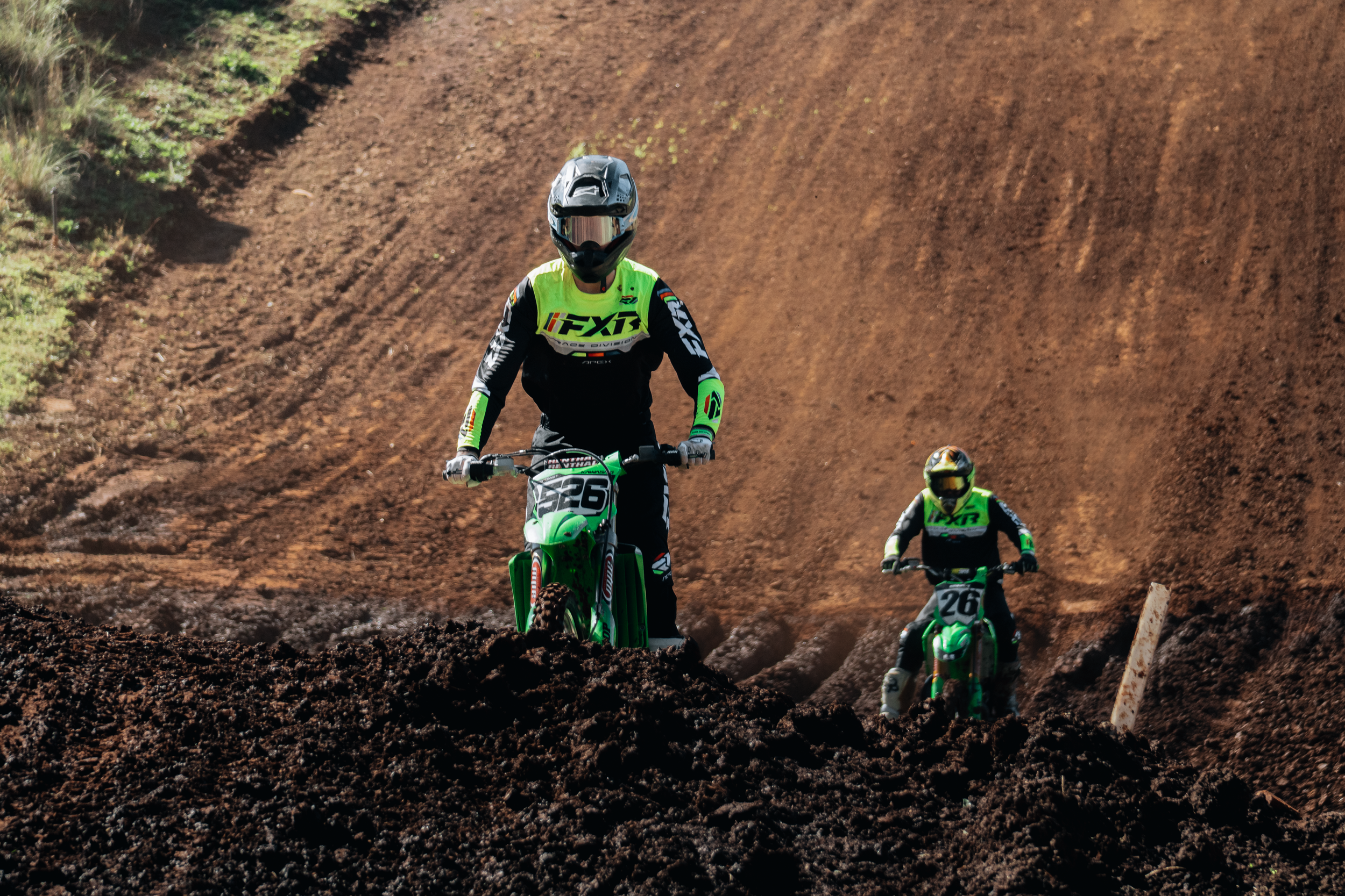Three motocross riders wearing helmets and bright green and black gear riding dirt bikes on a dirt track with a steep incline.