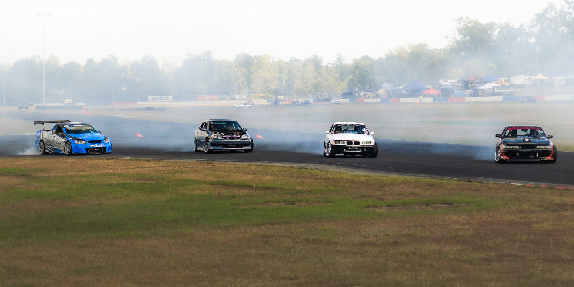 Four race cars on a racetrack creating tire smoke as they drift during a race event, with spectators and tents in the background.