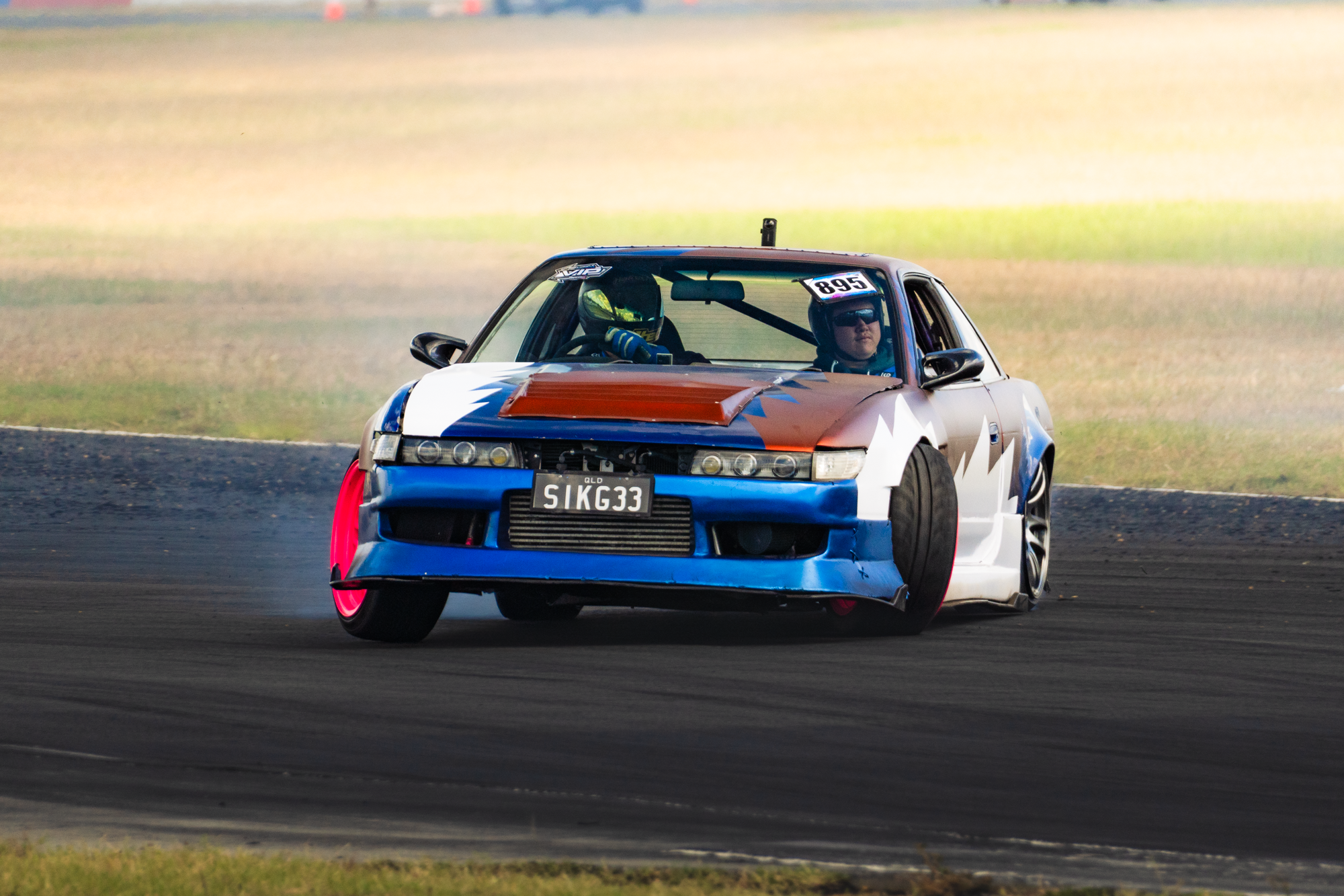 A race car with a colorful paint job, number 995, and a black windshield, driving on a race track with grass and blurred background.