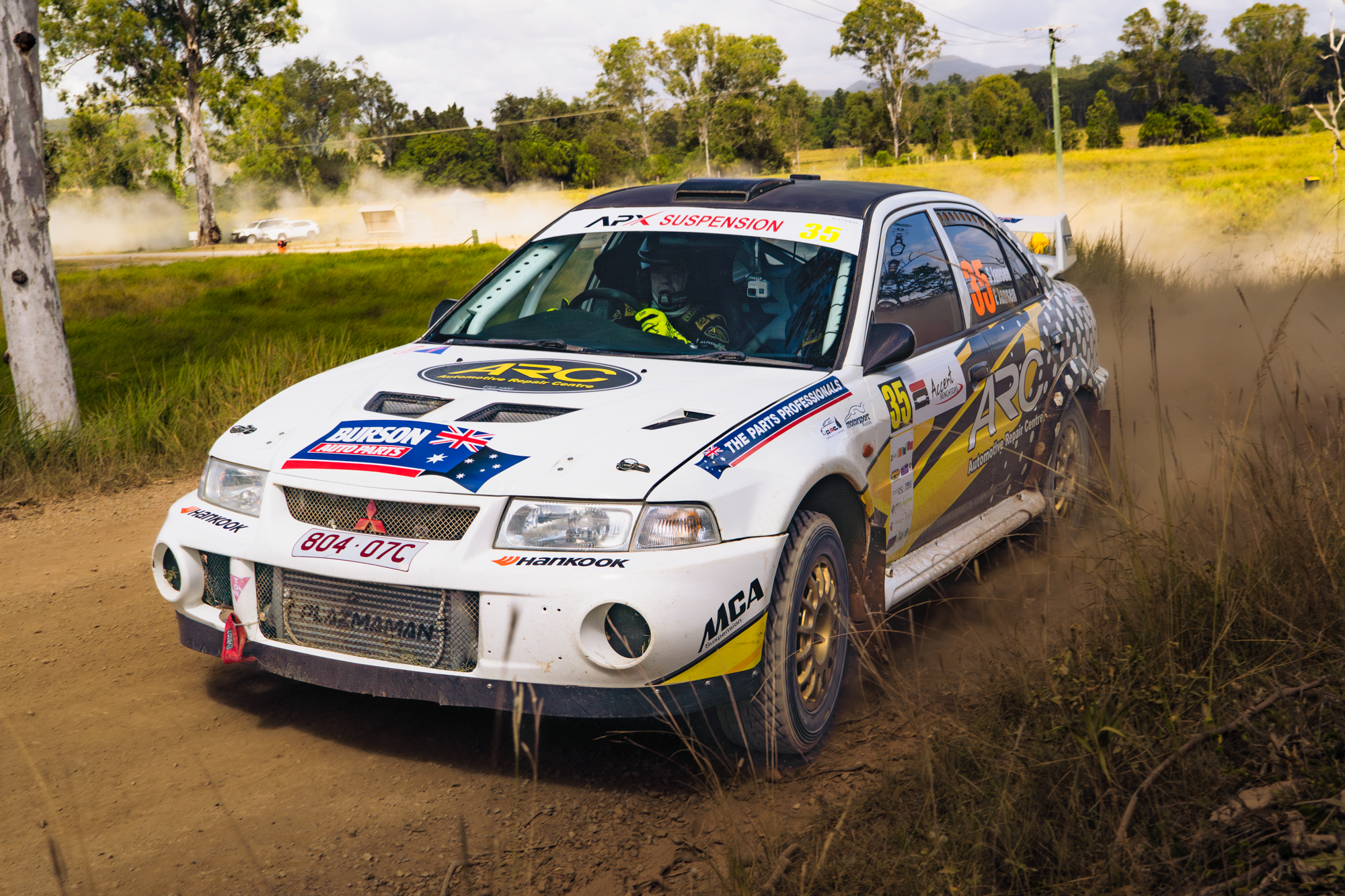 A white rally car with various sponsor logos speeds along a dirt road, kicking up dust, surrounded by green grass and trees, with a backdrop of cloudy sky and distant hills.