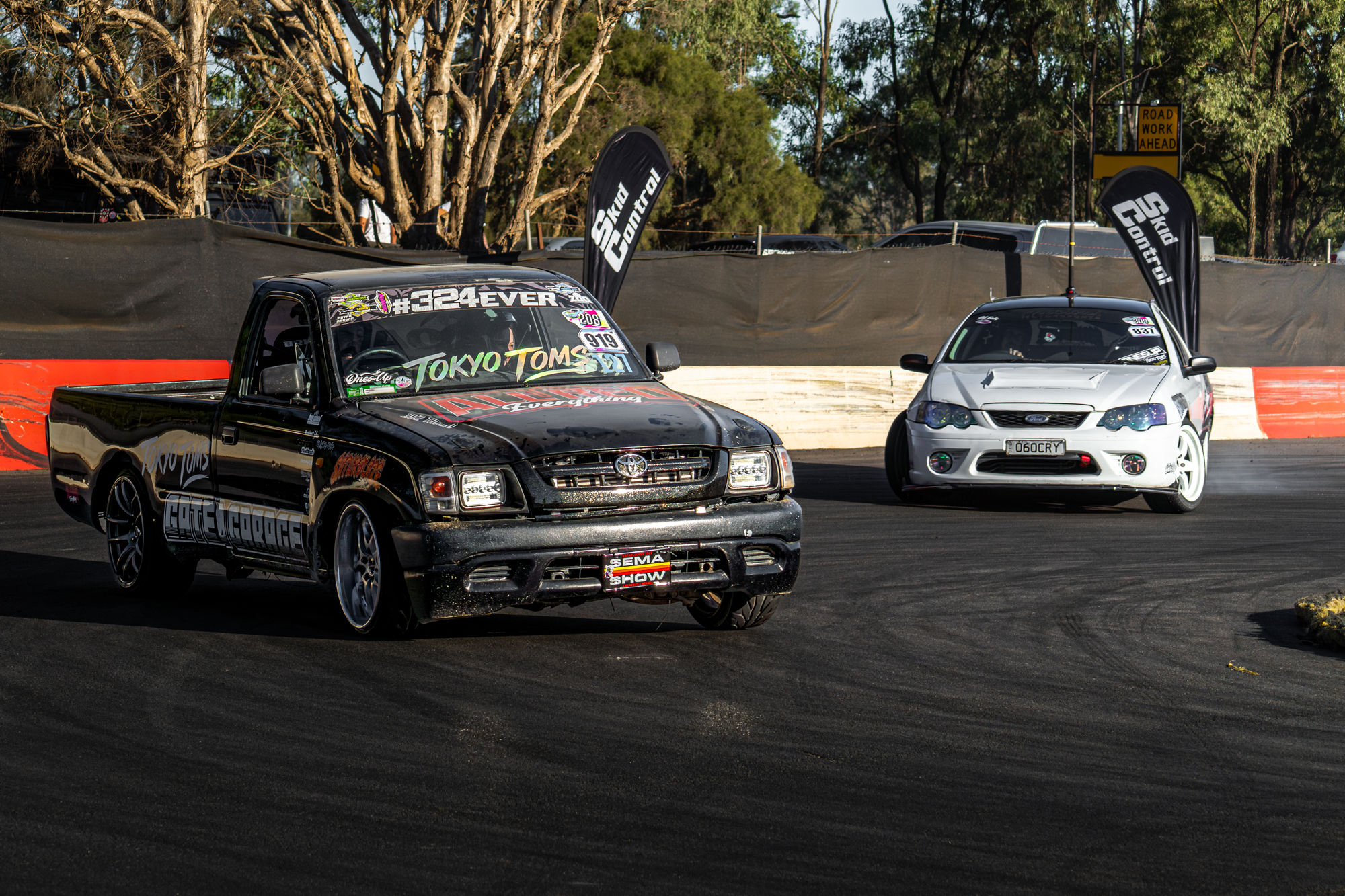 Two cars participating in a motorsport event on a race track, with flags and trees in the background, some racing banners, and a sign indicating road work ahead.