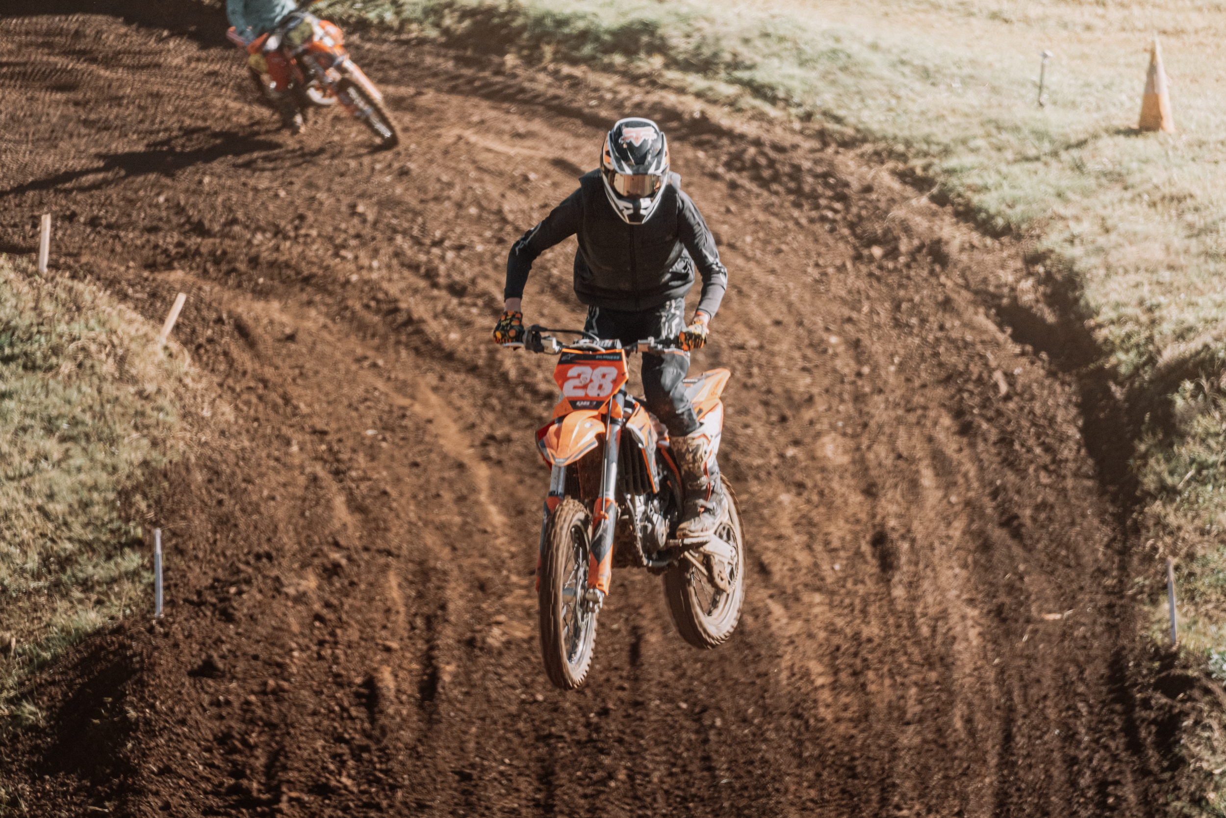 A person riding an orange dirt bike on a dirt track, wearing a black helmet and all-black clothing, with another rider in the background.