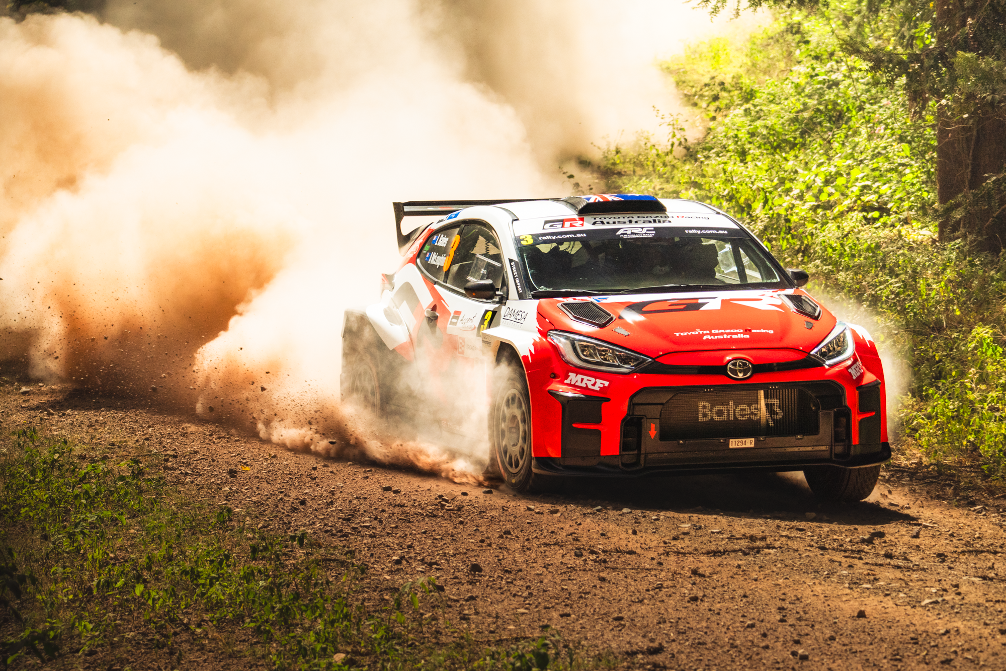 A red and white Toyota Gazoo Racing rally car driving on a dirt road, kicking up dust and gravel, surrounded by green foliage.