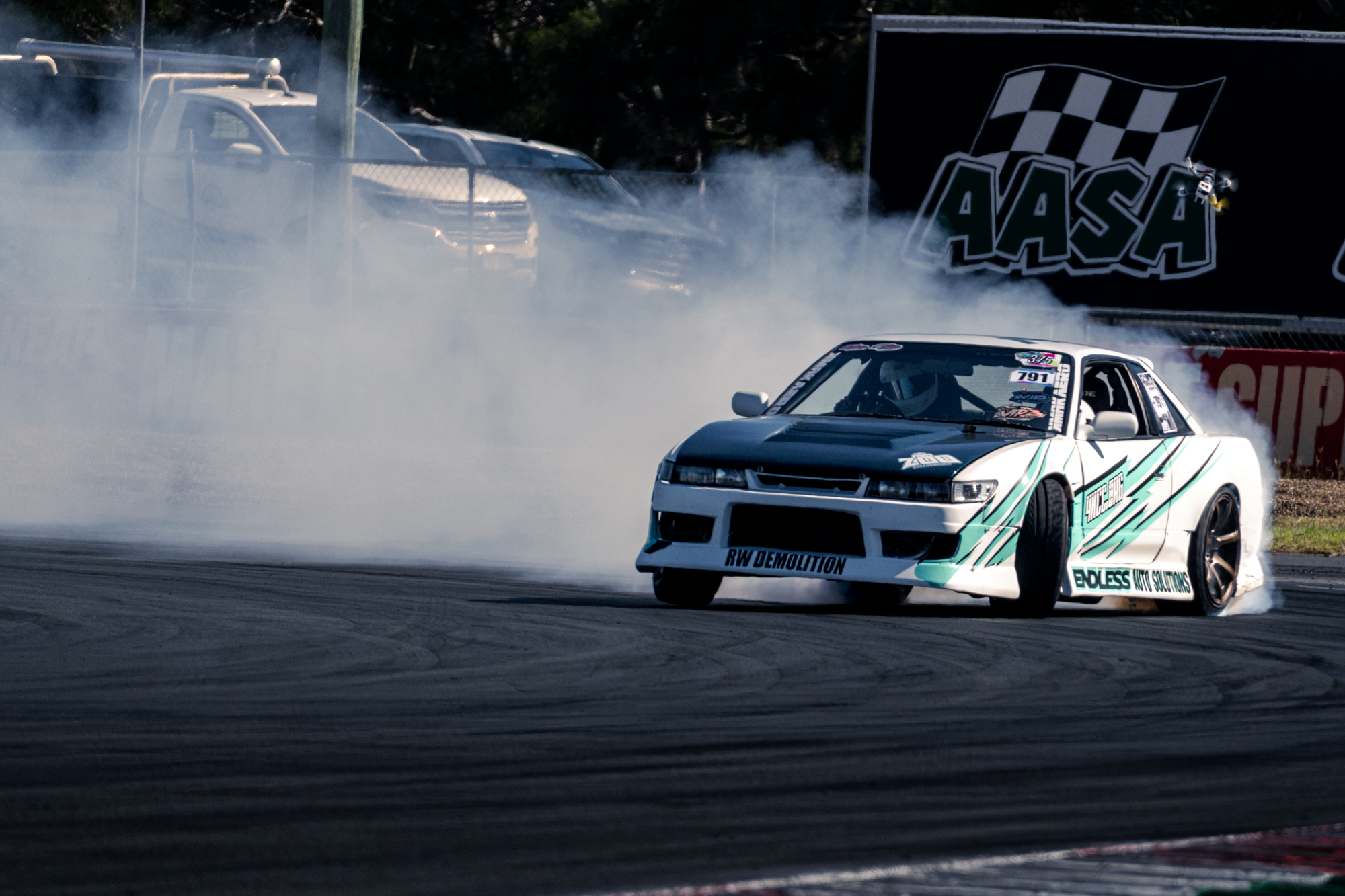 A silver and teal race car drifting on a track, producing tire smoke, with a background of vehicles and a black banner that reads 'VASA' and features a checkered flag logo.