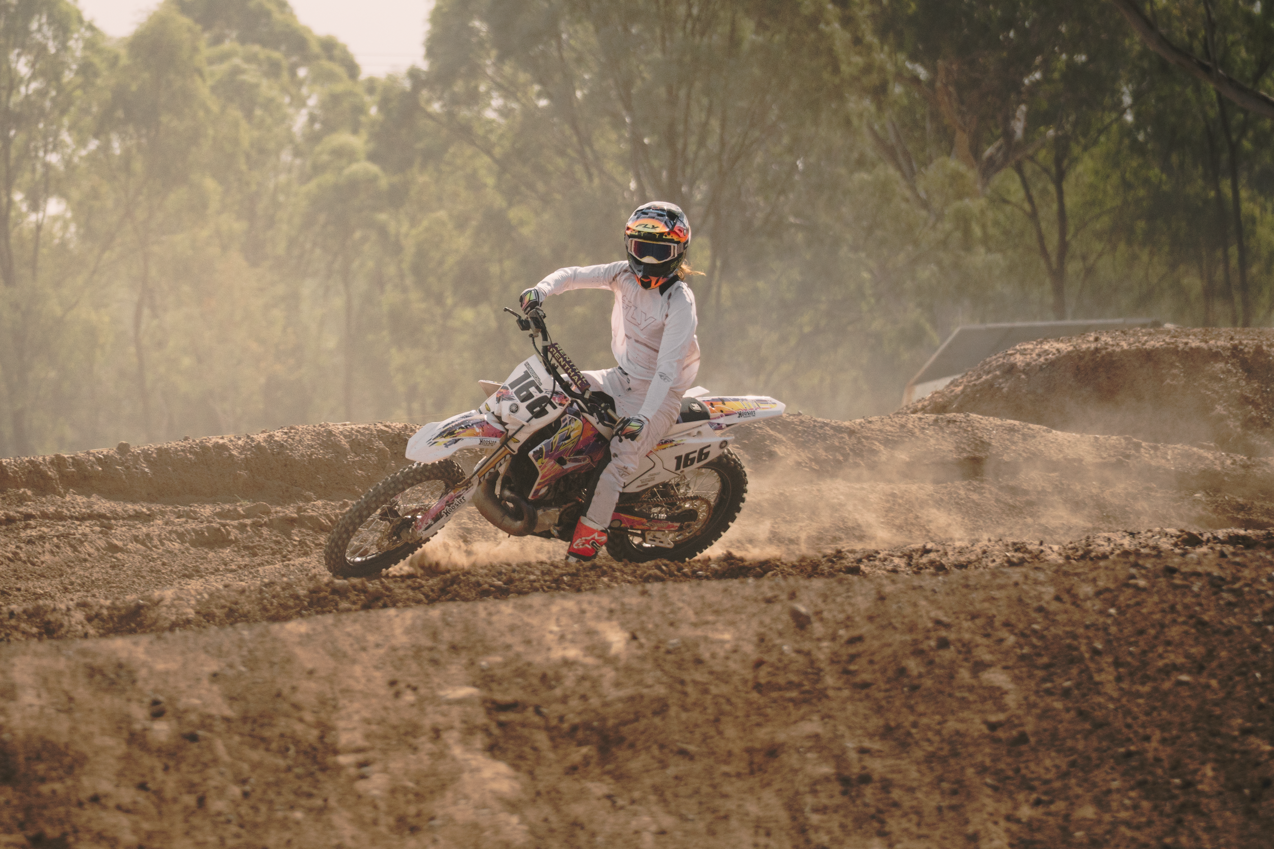 A person wearing a helmet and protective gear riding a dirt bike on a dusty track with a forested background.