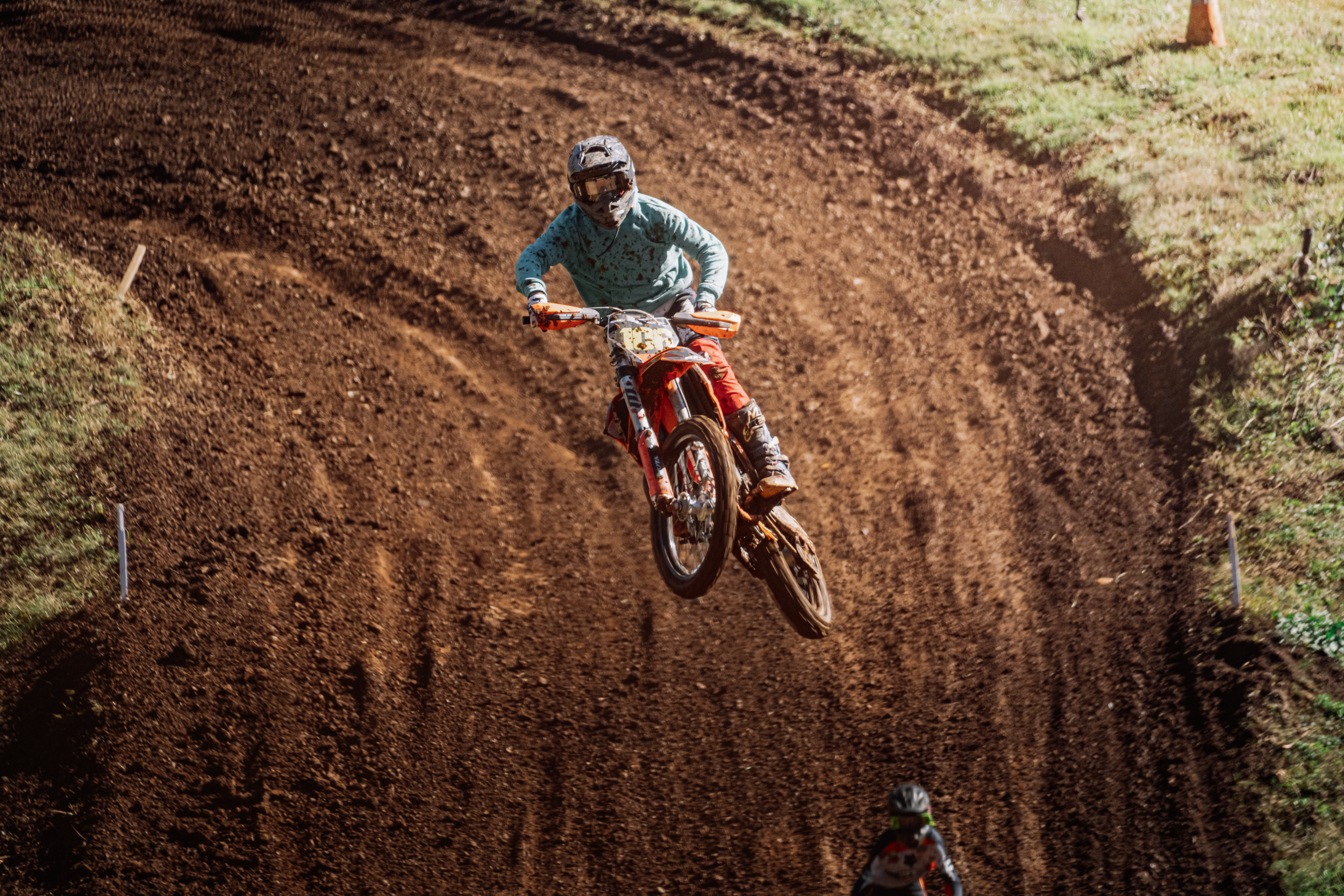 A person riding a dirt bike on a motocross track, wearing a helmet and riding gear, with a dirt trail and grass on the sides.