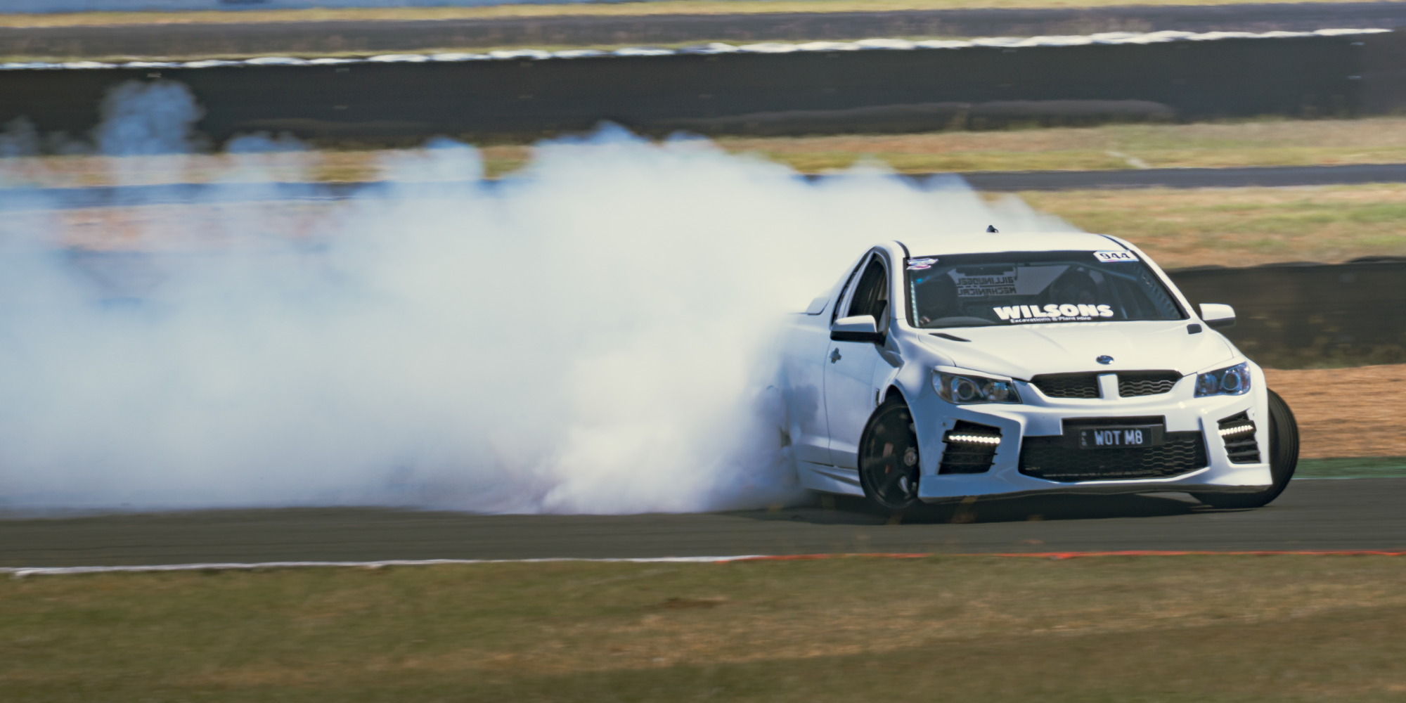 A white sports car drifting on a race track, creating smoke from its tires with a blurred background of grass and asphalt.
