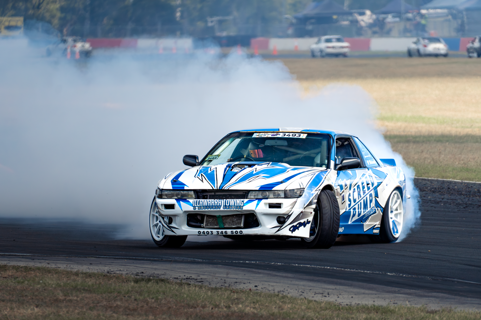 A blue and white sports car with aftermarket modifications performs a drifting maneuver on a race track, creating tire smoke. The car displays sponsorship logos, including 'Illawarra Towing & Recovery Service,' and has a racing number '3493' on the w