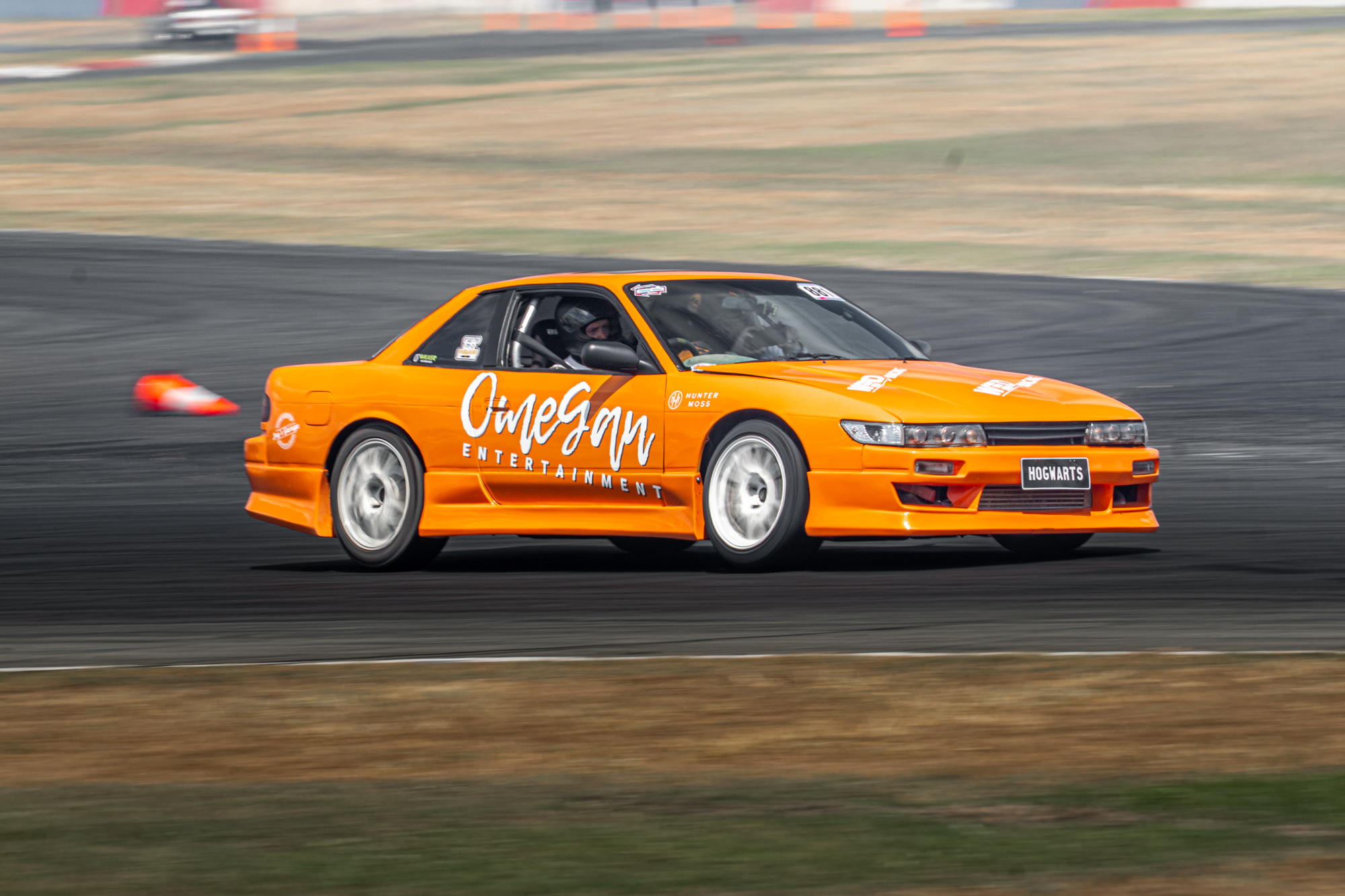 An orange racing car with 'Omega Entertainment' written on the side, driving on a racetrack with motion blur, and a driver inside wearing a helmet.