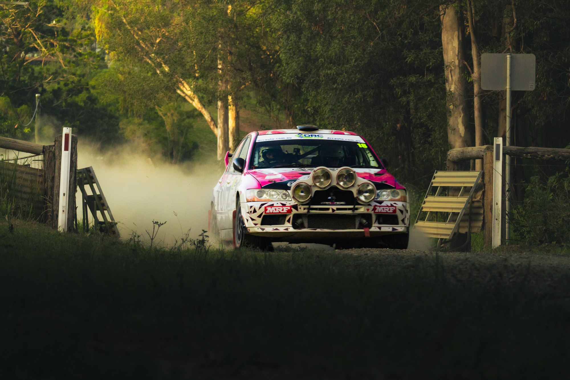 A rally car with multiple large front lights on its front bumper driving on a dirt road through a wooded area, kicking up dust.