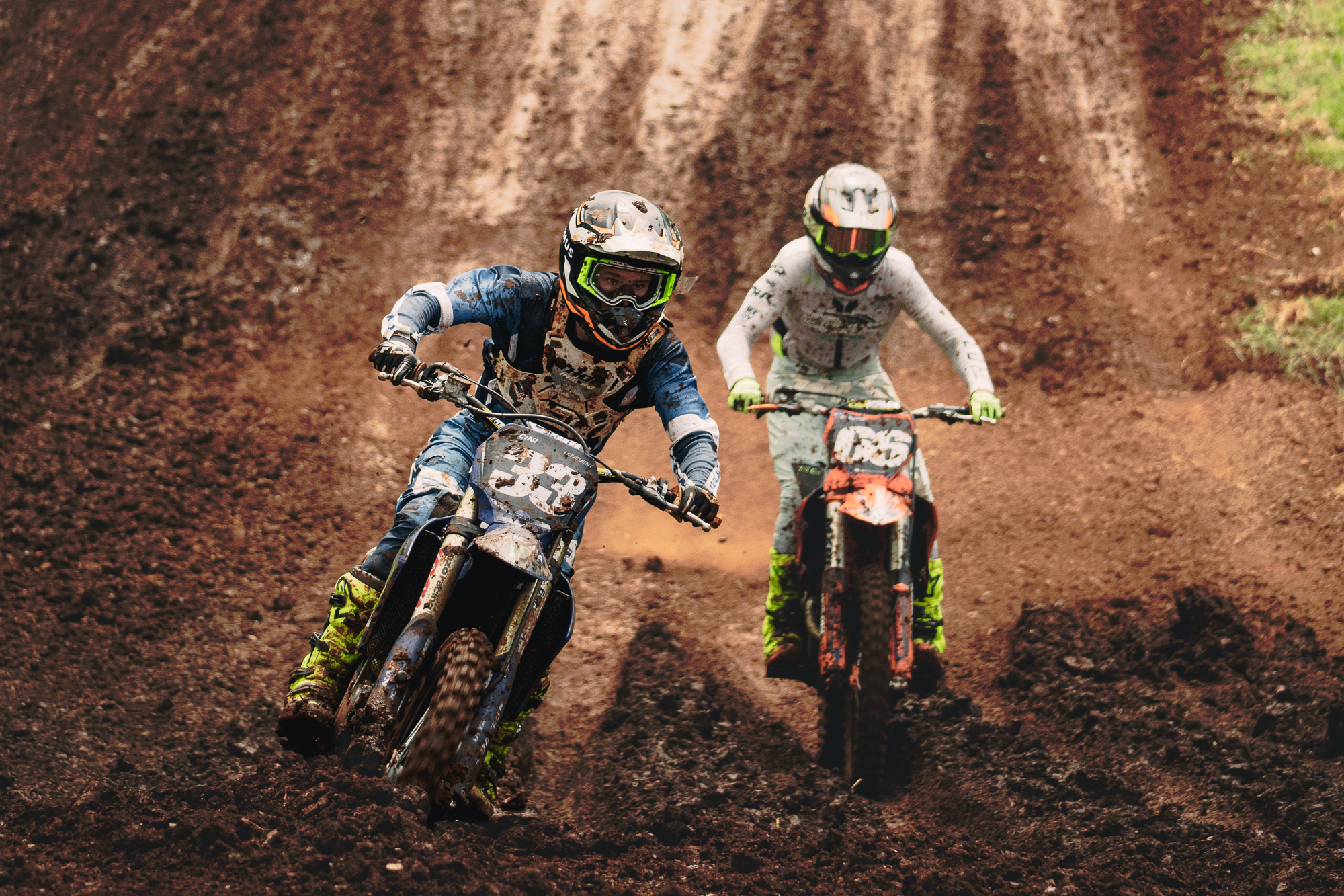 Two motocross riders dressed in protective gear and helmets riding dirt bikes on a muddy off-road track.