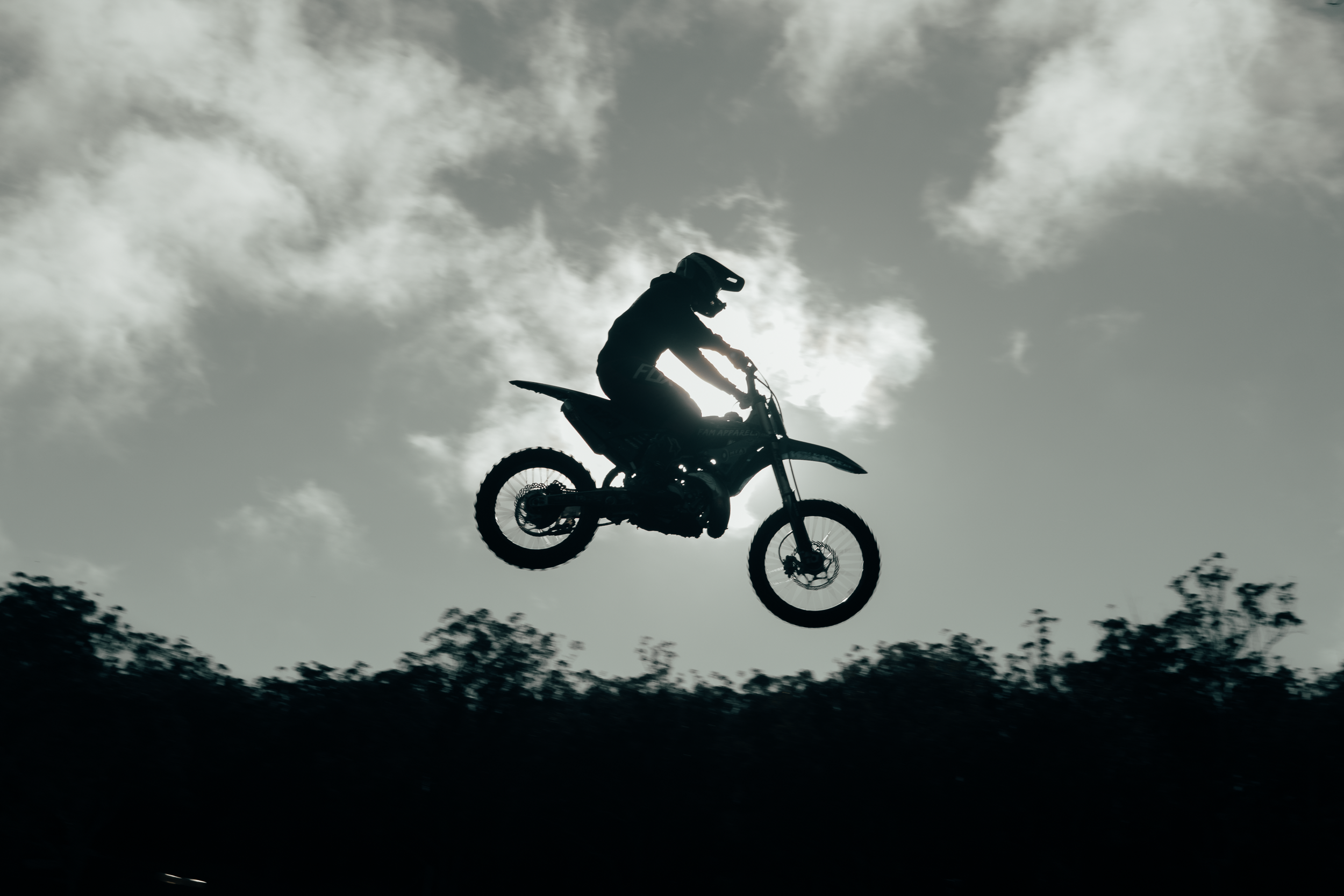Silhouette of a person riding a dirt bike in mid-air against a cloudy sky with the sun shining through.