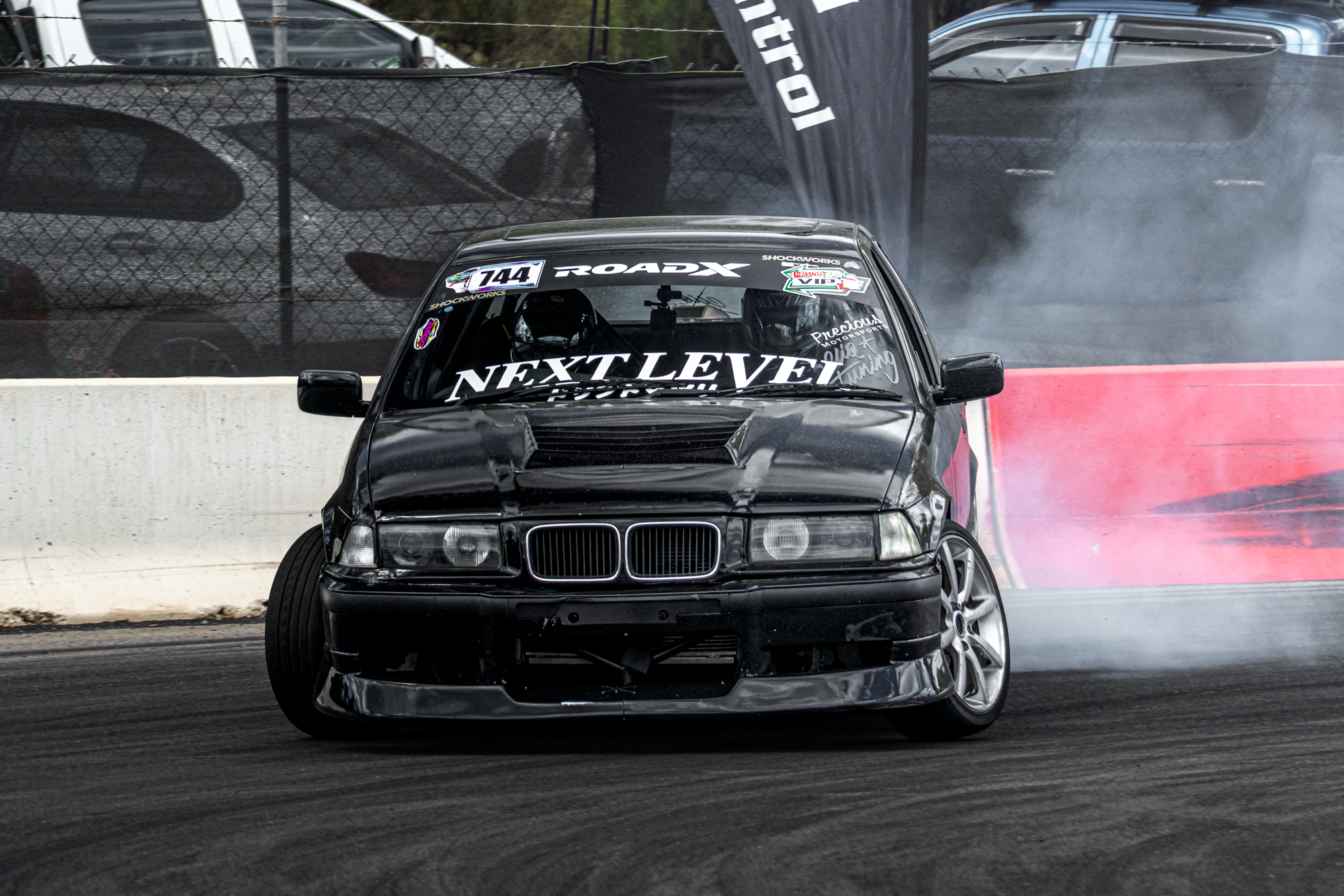 A black BMW race car with racing decals, including 'NEXT LEVEL' on the windshield, is drifting on a race track, creating smoke from the tires while near the track's concrete barrier.