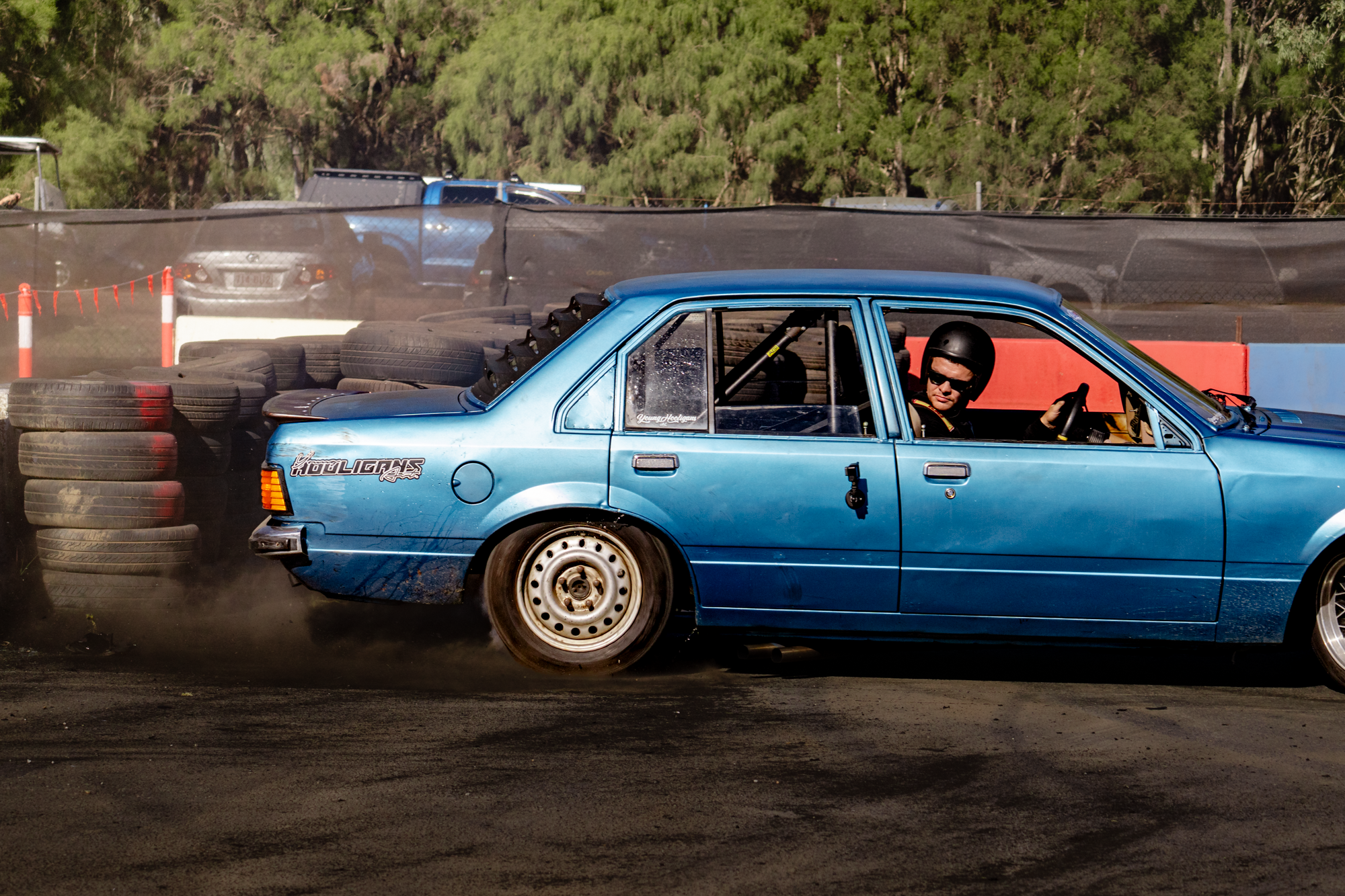 A blue car with a modified rear end and racing stickers is involved in a burn-out during a racing event. The car's rear tires are spinning and creating smoke, with a driver wearing a black helmet giving a thumbs-up. Stacked tires and other vehicles c