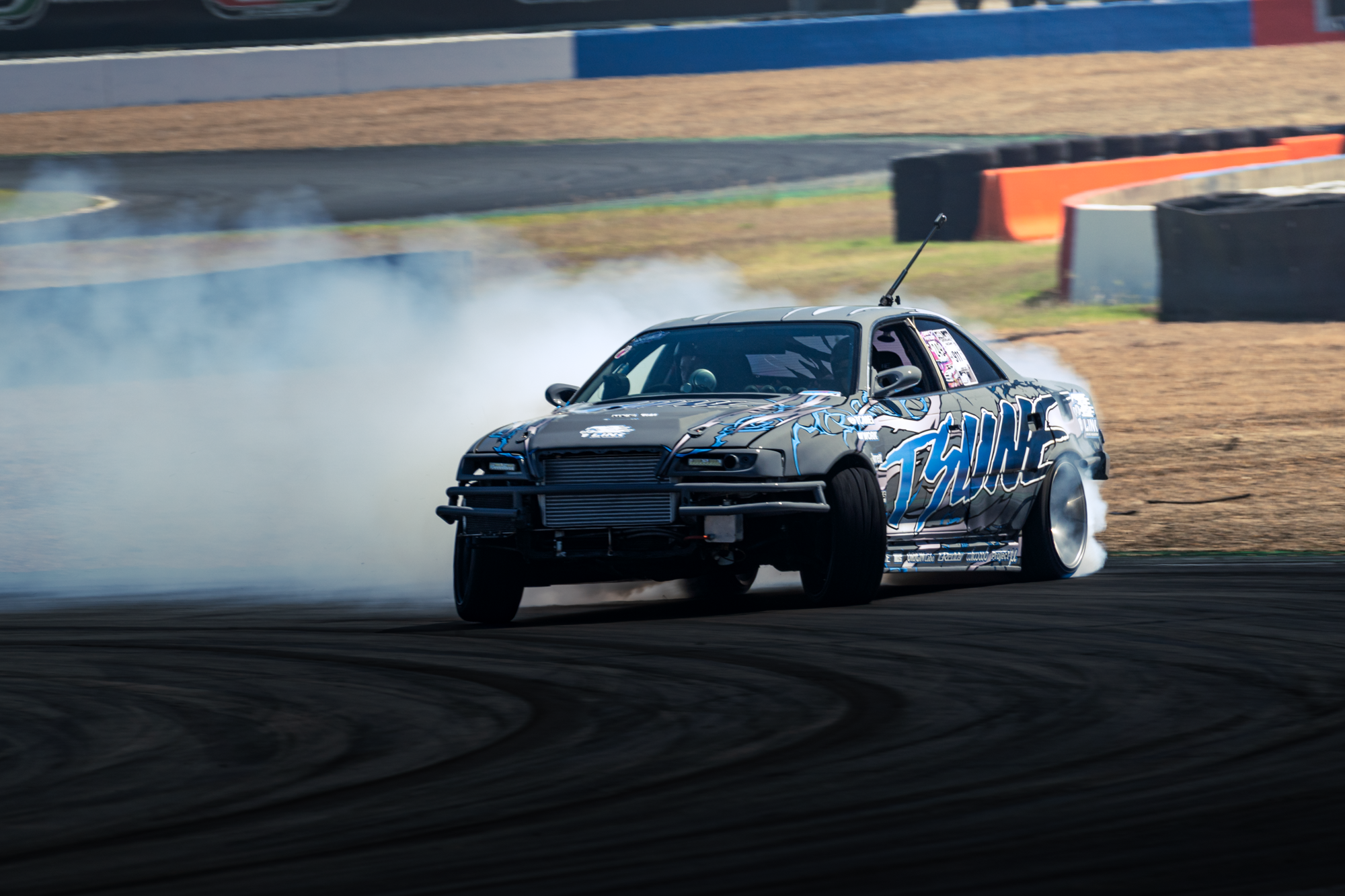 A black sports car with blue and white decals drifting on a racetrack, producing smoke from the tires, with barriers on the side of the track.
