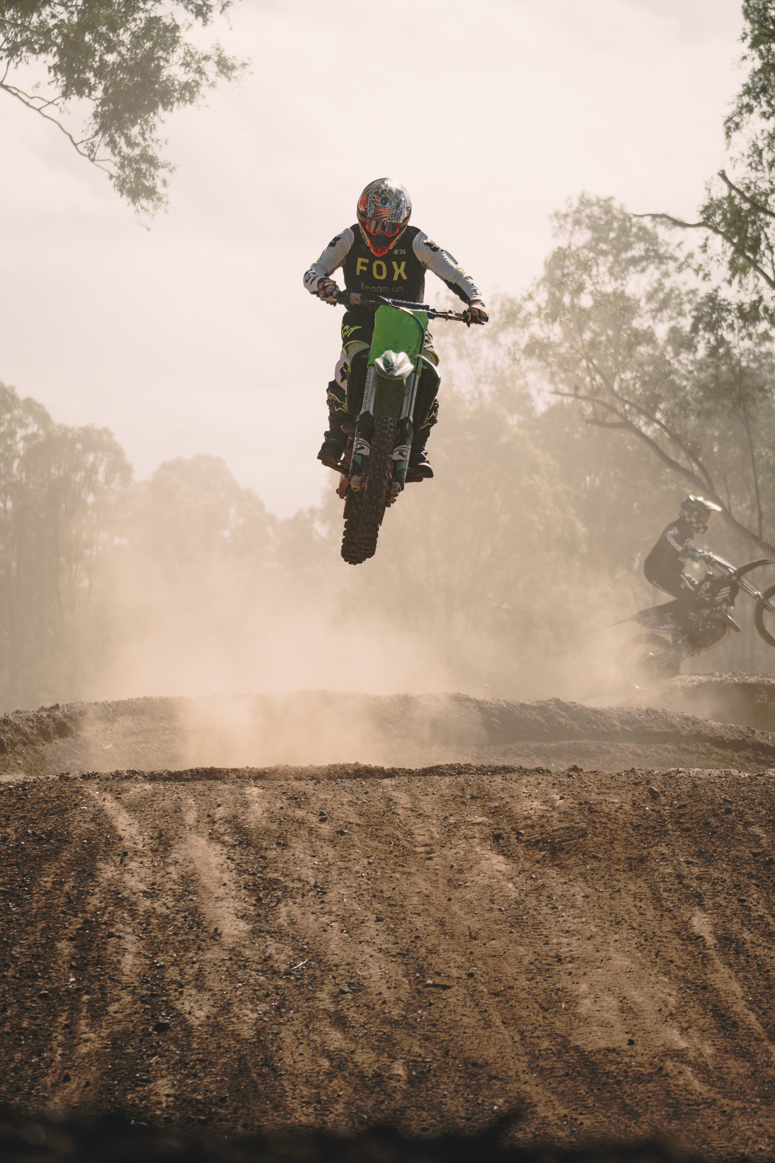 A motocross rider airborne above a dirt track wearing a helmet and protective gear, with a second rider visible in the background and dust clouds behind them.