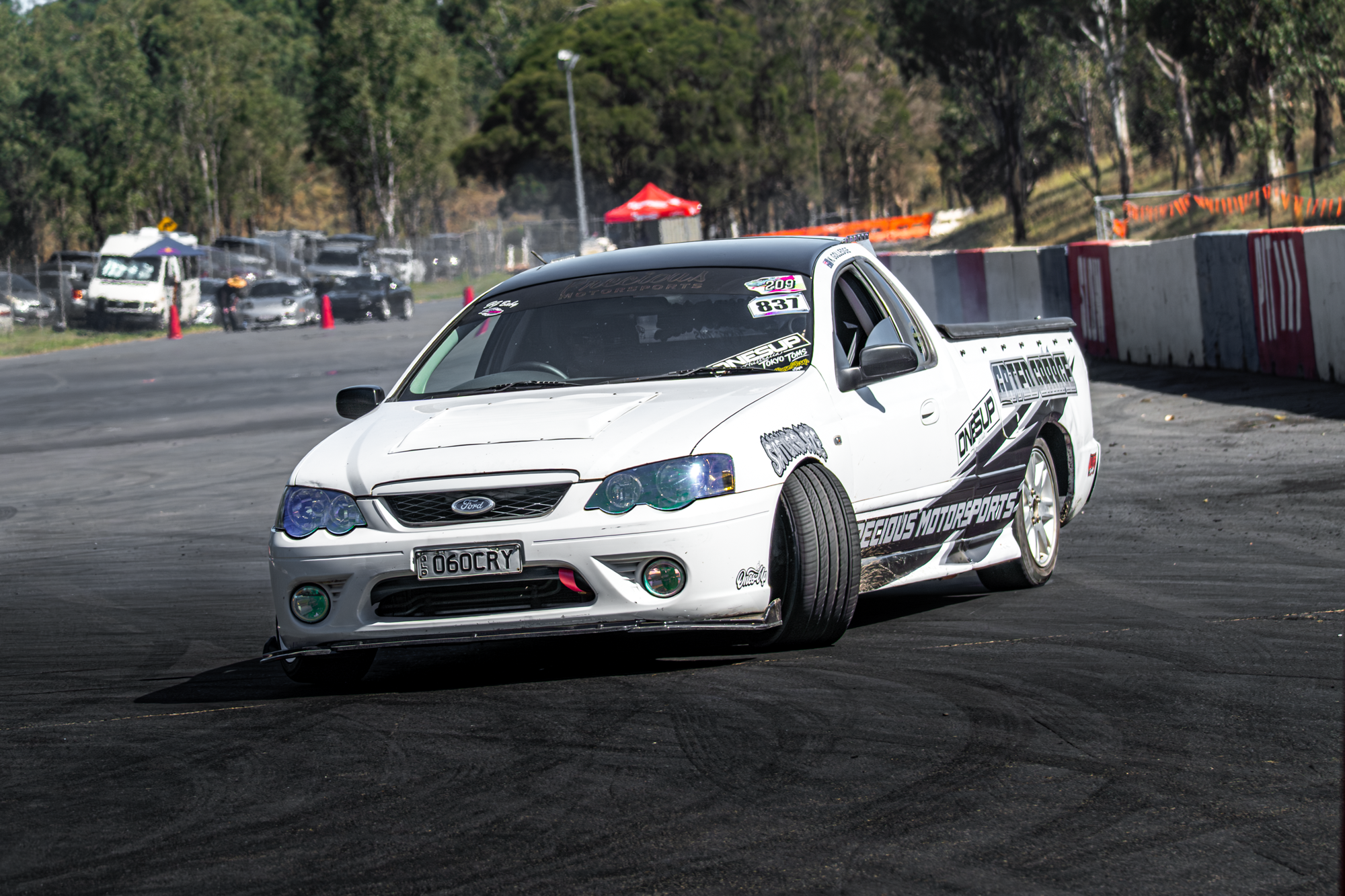 White race car with racing decals driving on a track, with a line of cars and trees in the background.