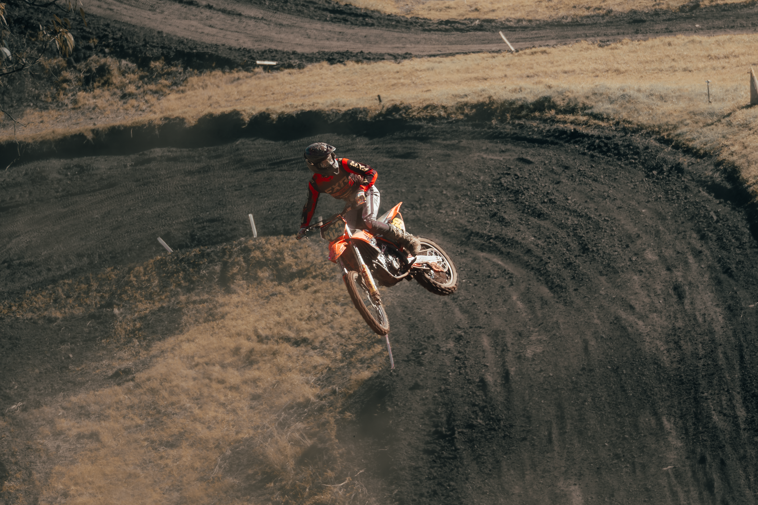 A motocross rider in red gear riding on a dirt track, captured mid-air during a jump.