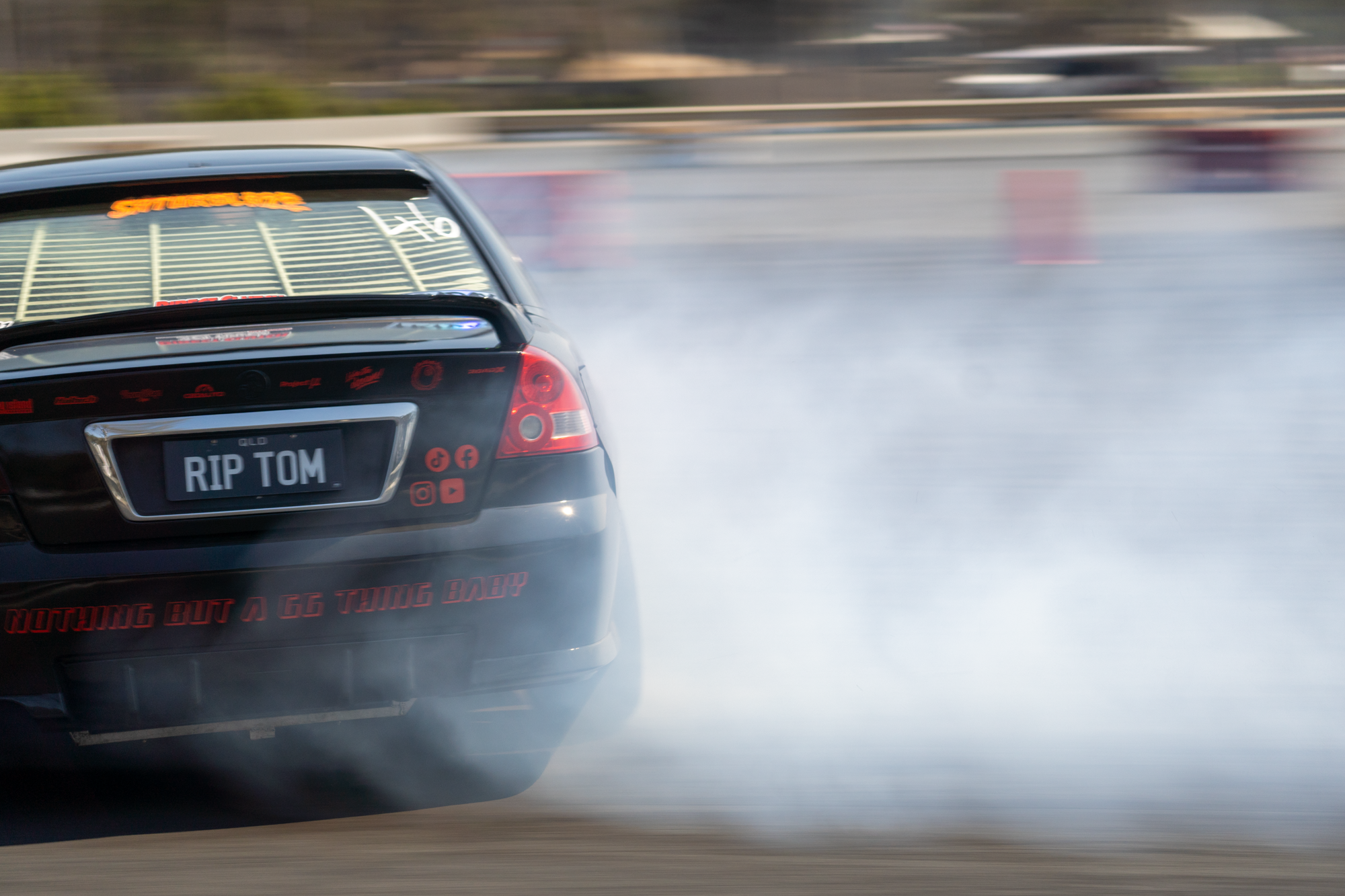 A black car performing a burnout on a race track, with smoke billowing from the tires, and a license plate that reads 'RIP TOM,' with various stickers and decals on the rear window and bumper.