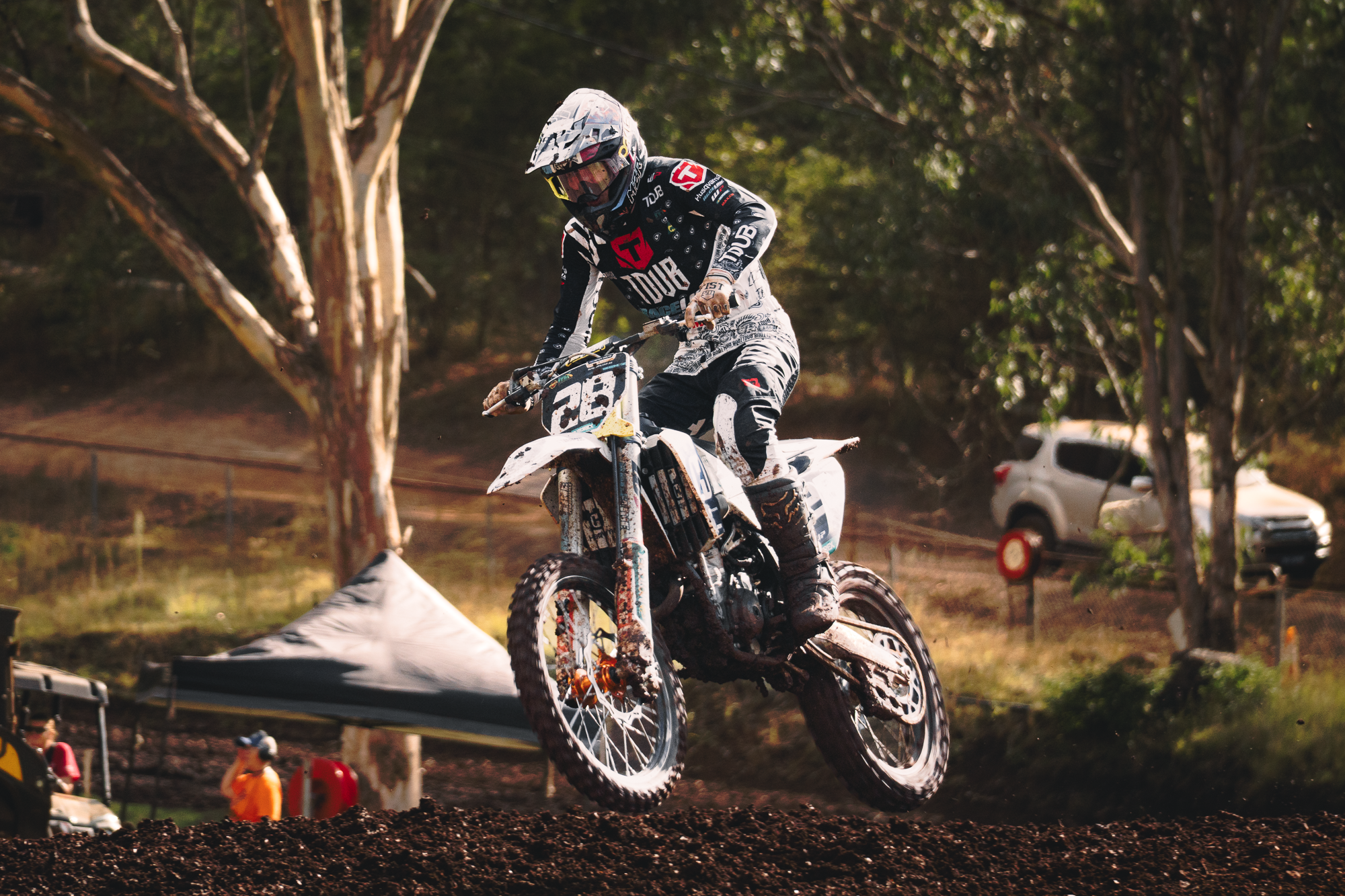 A motocross rider in black and white gear, wearing a helmet and goggles, riding a dirt bike through a dirt track surrounded by trees and spectators.