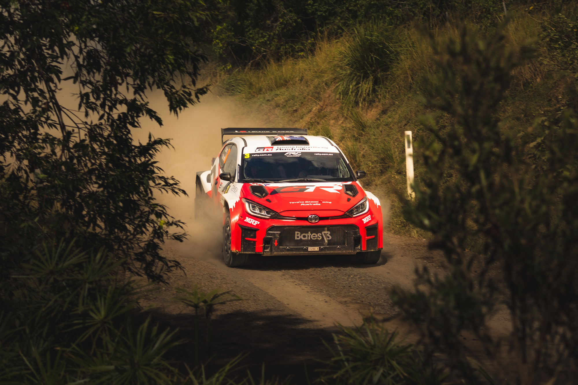 A rally race car driving on a dirt road through a wooded area, kicking up dust, with lush green foliage on both sides.