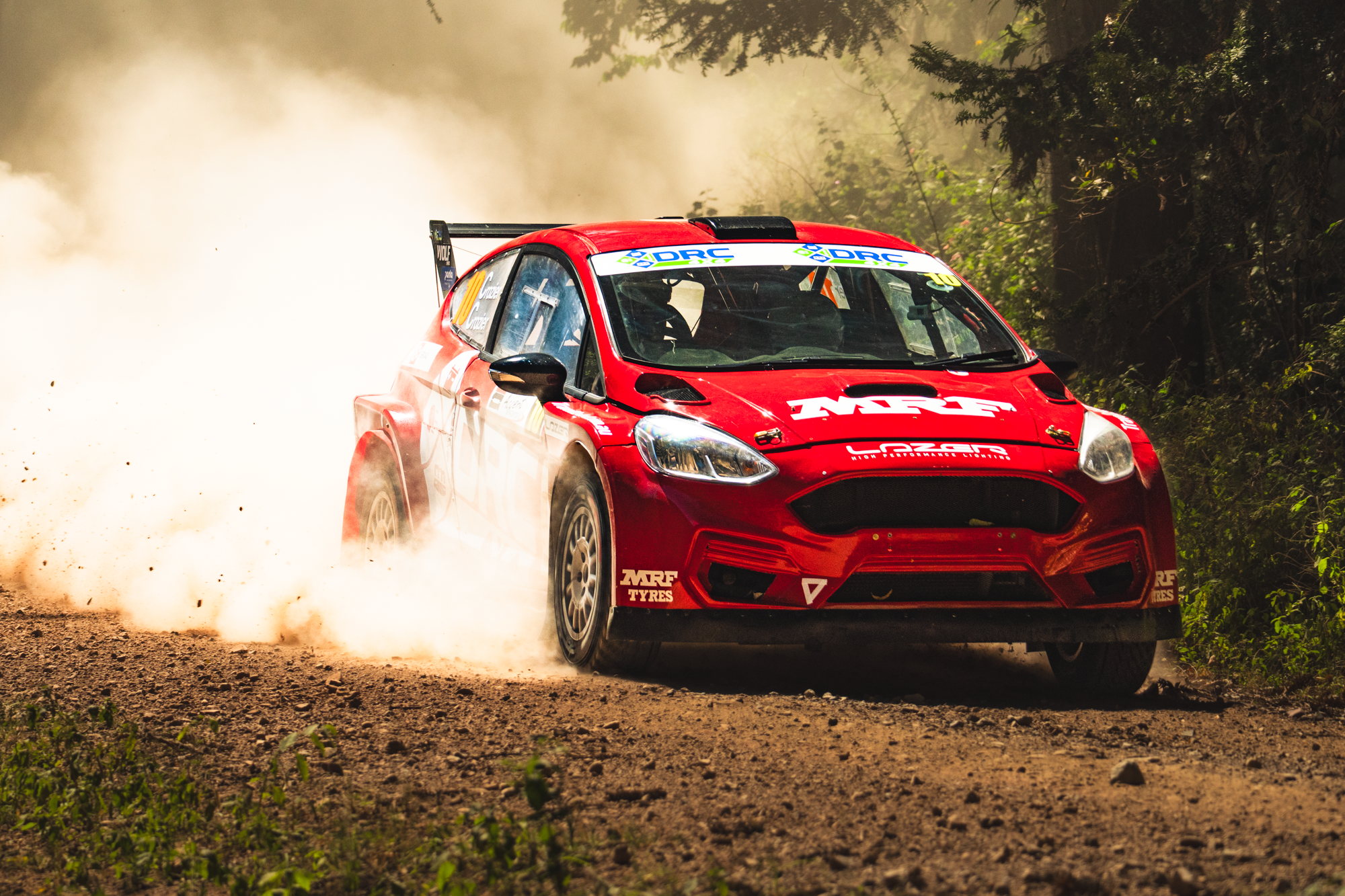 A red rally car kicking up dust and dirt on a gravel dirt road in a forested area during a race.