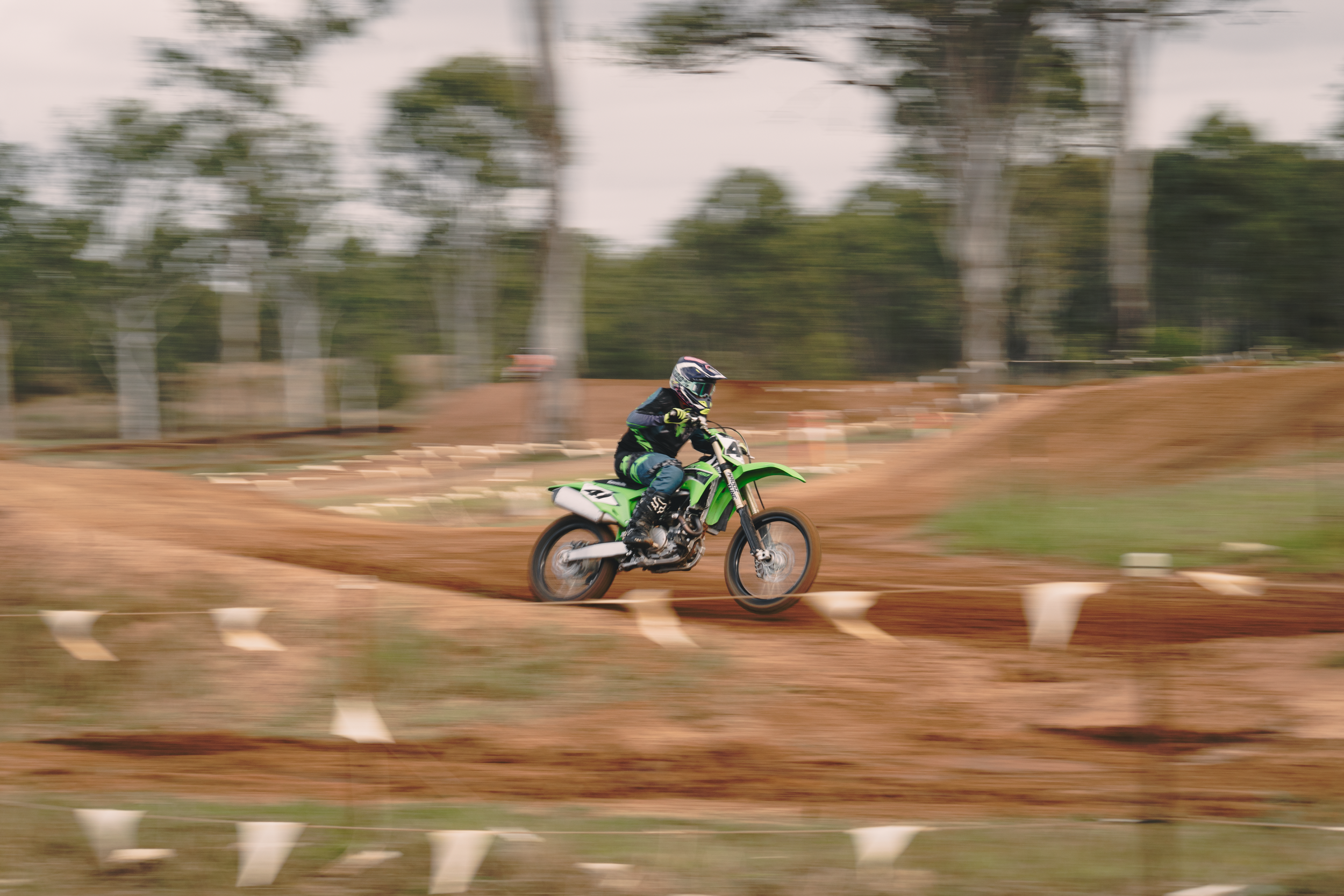 A person riding a green dirt bike on a dirt track with trees in the background, captured with motion blur.