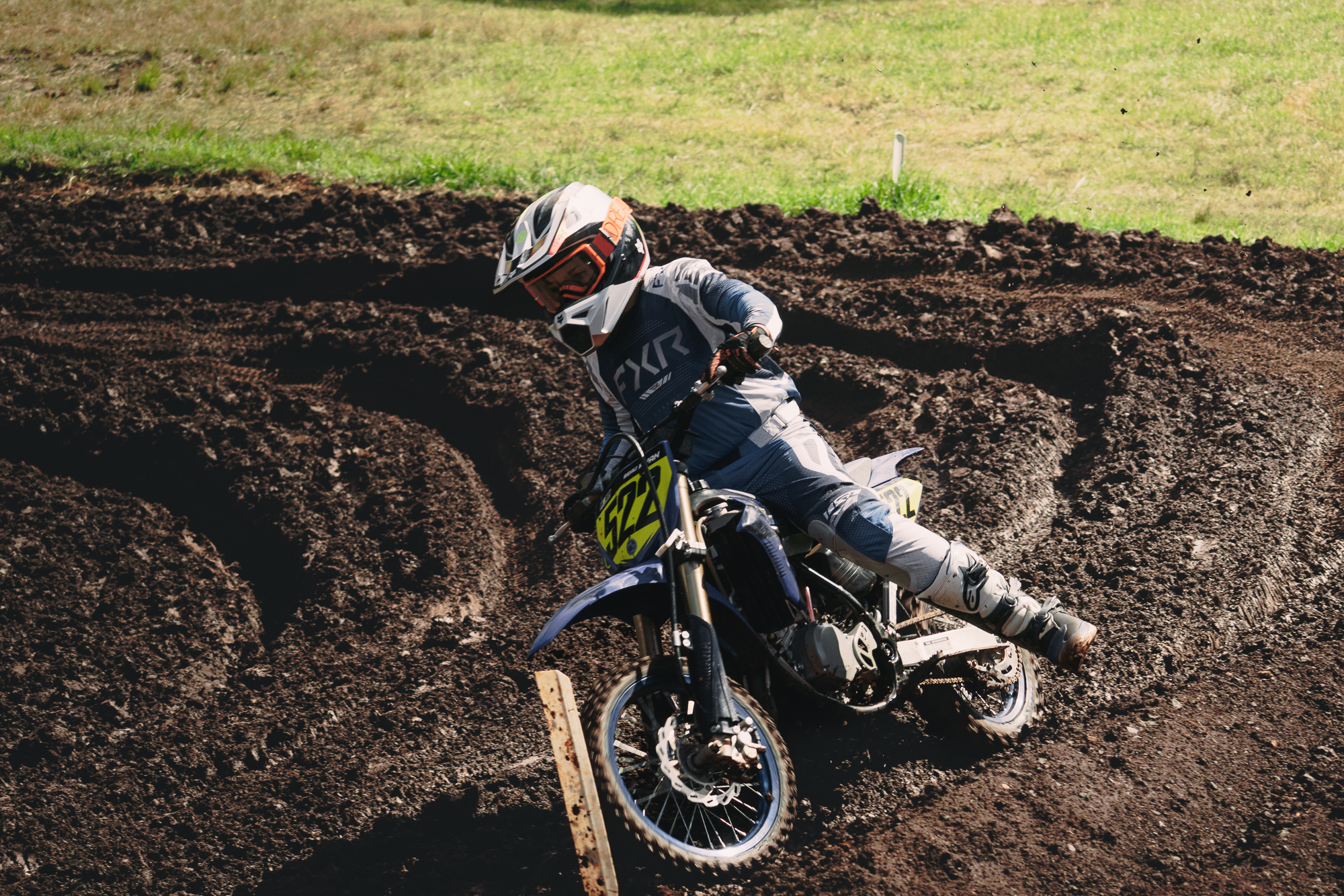 Motocross rider wearing a helmet and racing gear, leaning into a turn on a dirt track on a sunny day.
