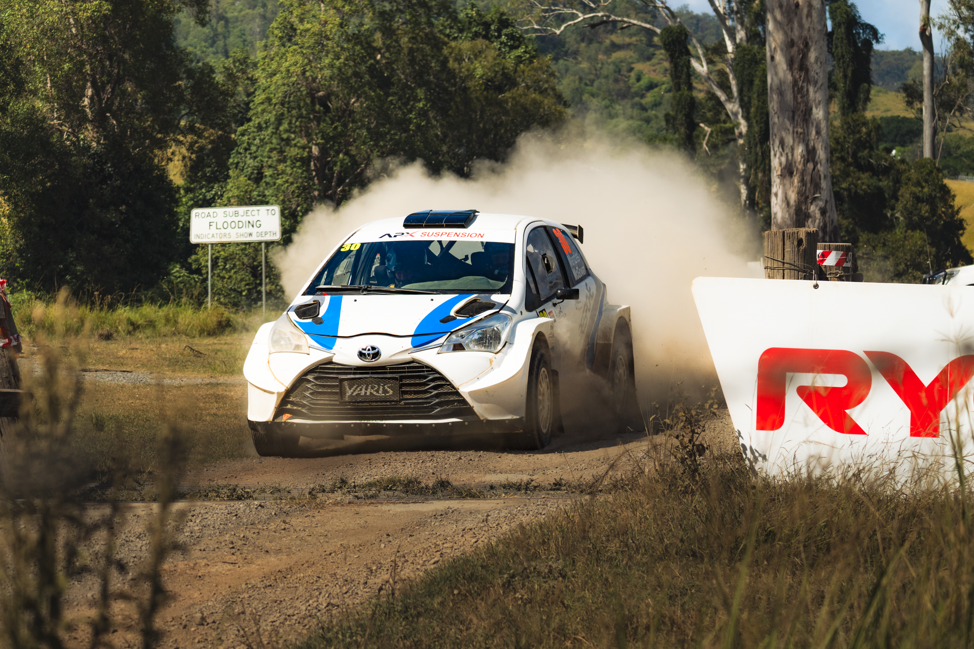 A white rally car with blue accents on a dirt race track, kicking up dust as it navigates a turn. The car is marked with the number 30 and sponsored by APX Suspension. A sign in the background warns of flooding on the road.