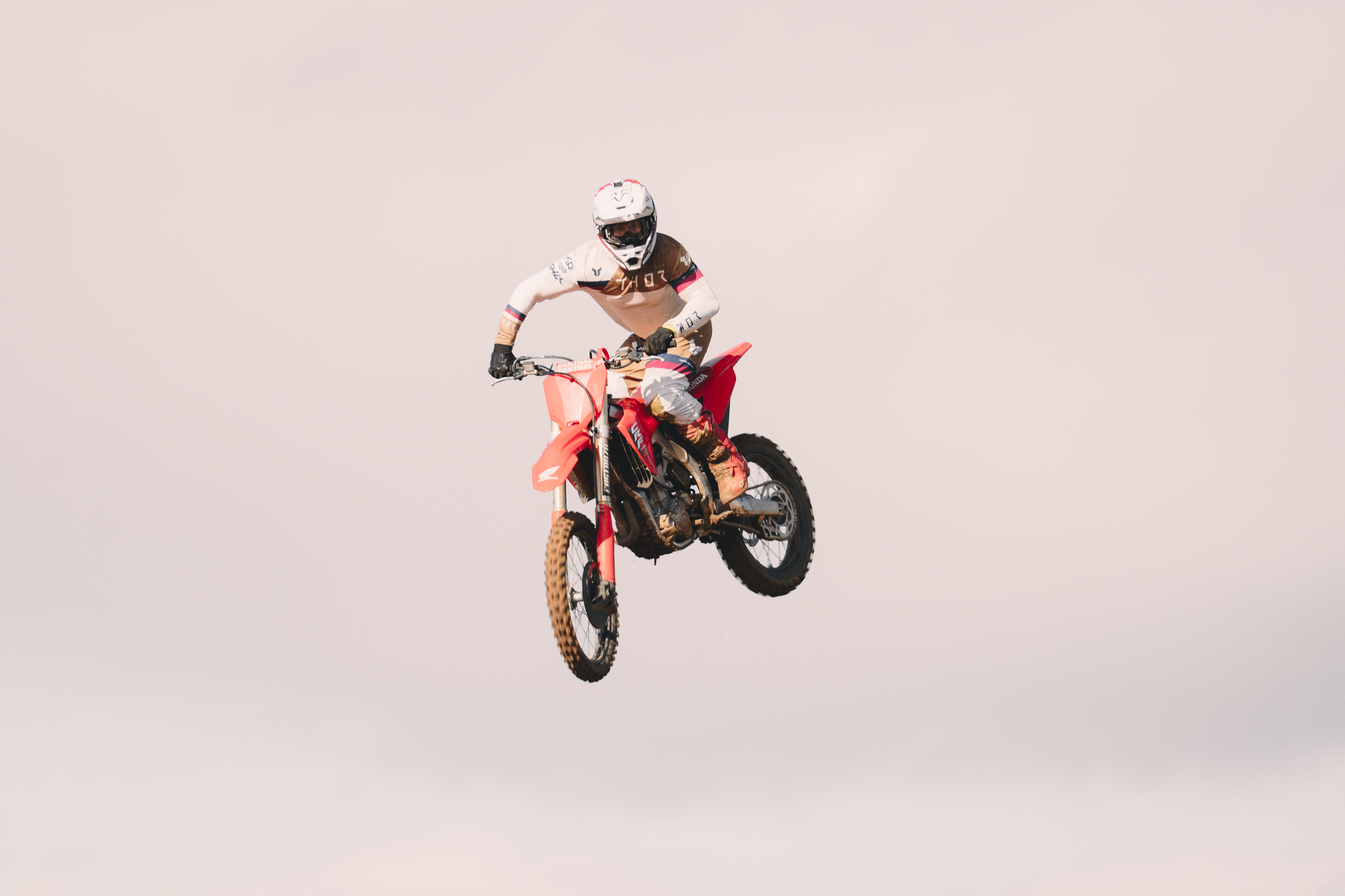 A person wearing full motocross gear, including a helmet and goggles, riding a red dirt bike mid-air against a plain sky background.