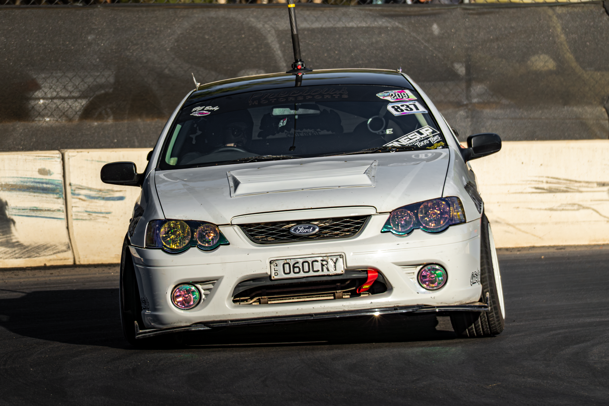 A white Ford race car with colorful headlights and a custom license plate '060CRY', driving on a race track with barriers on the side, during a race event.