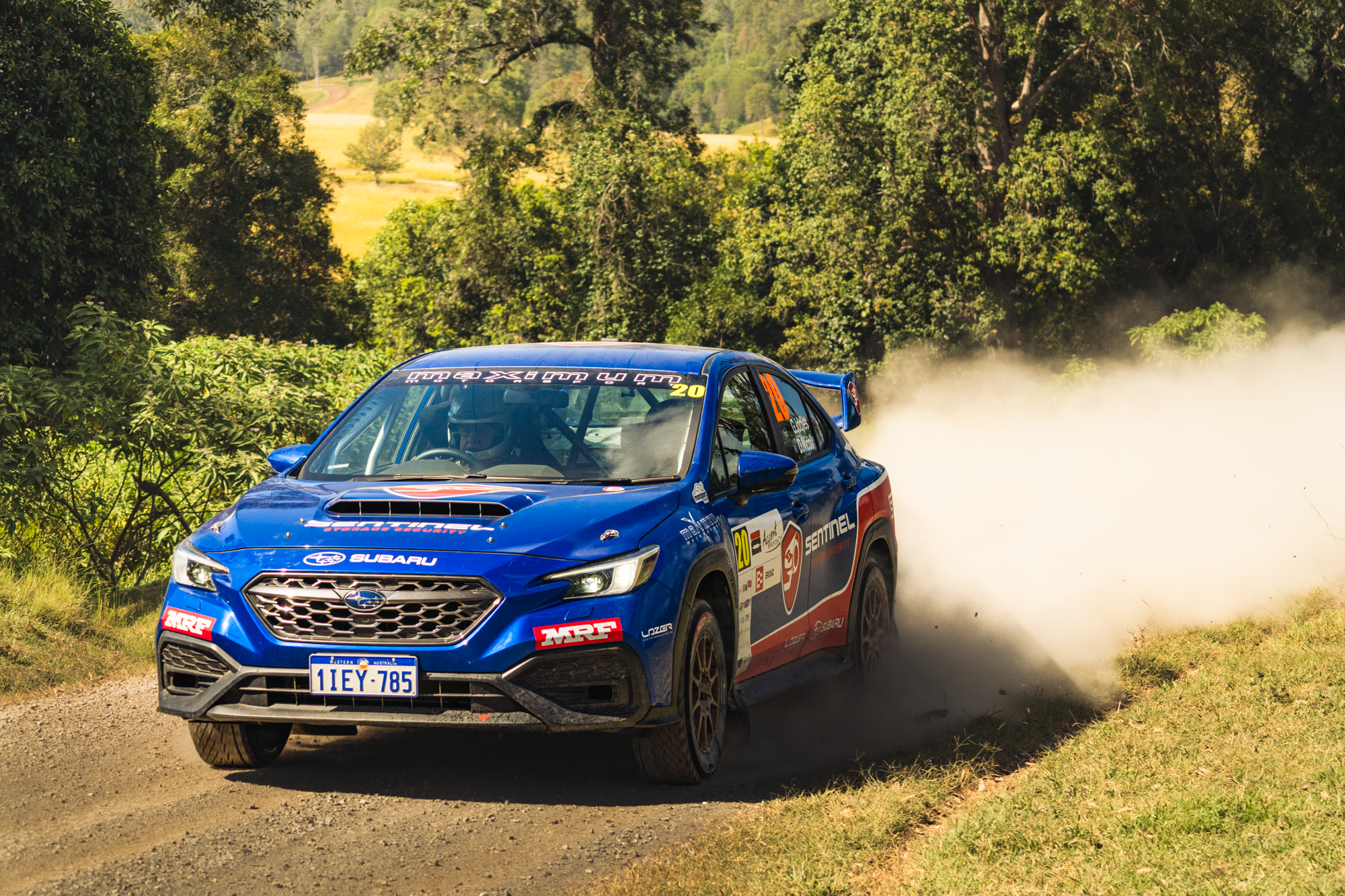 A blue Subaru rally car speeding on a dirt road, kicking up dust, surrounded by lush green trees and a rural landscape.