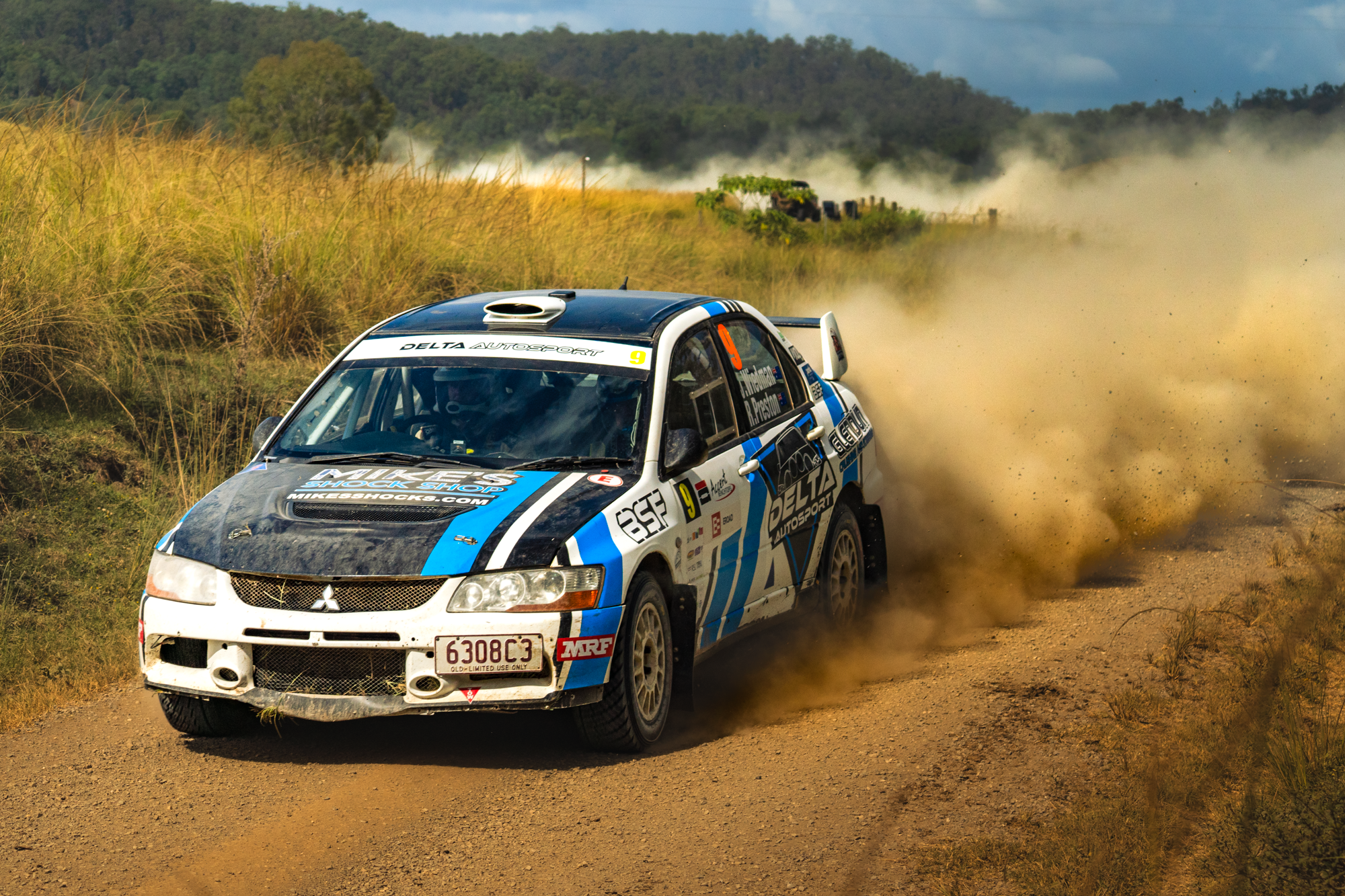 A rally race car speeding on a dirt road with dust clouds behind it, in a rural setting with grass and hills in the background.