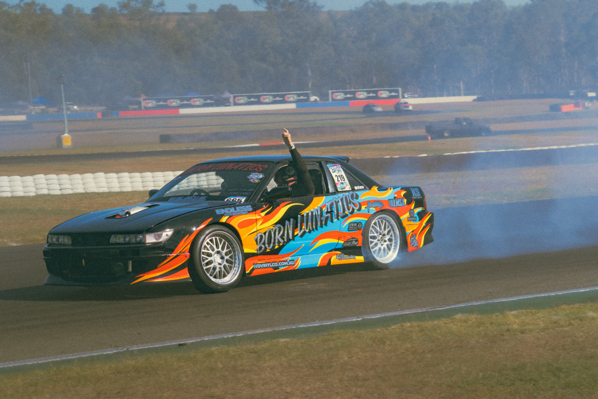 A race car with colorful flame decals, the word "Born-LUMATICS" on the side, drifting on a racetrack with smoke coming from the tires, a person inside raising their hand.