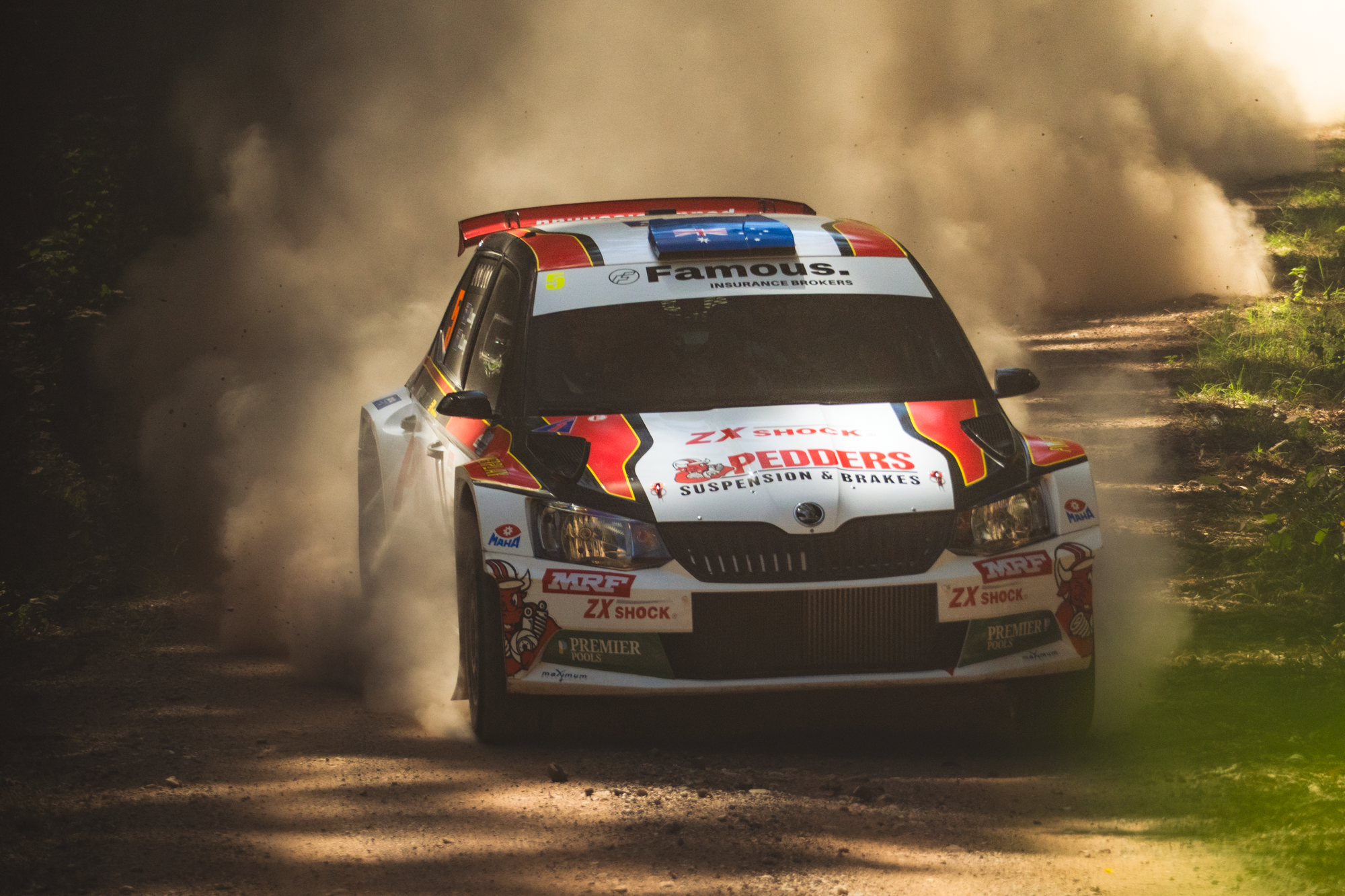 A rally car racing on a dusty dirt road through a wooded area, kicking up dust clouds behind it.