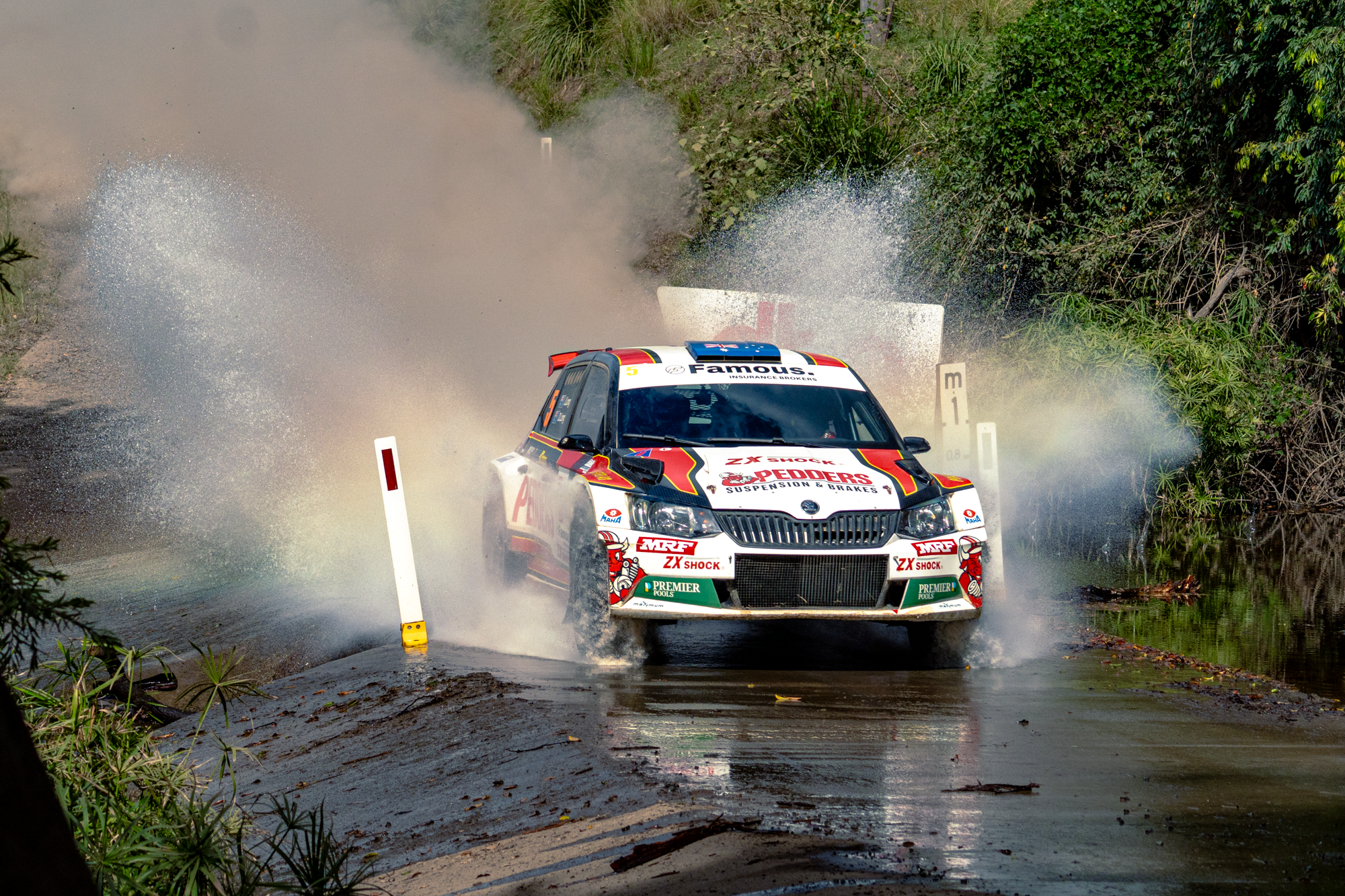 A rally car driving through water and mud on a dirt road surrounded by greenery, creating splashes while navigating a water crossing during a race.