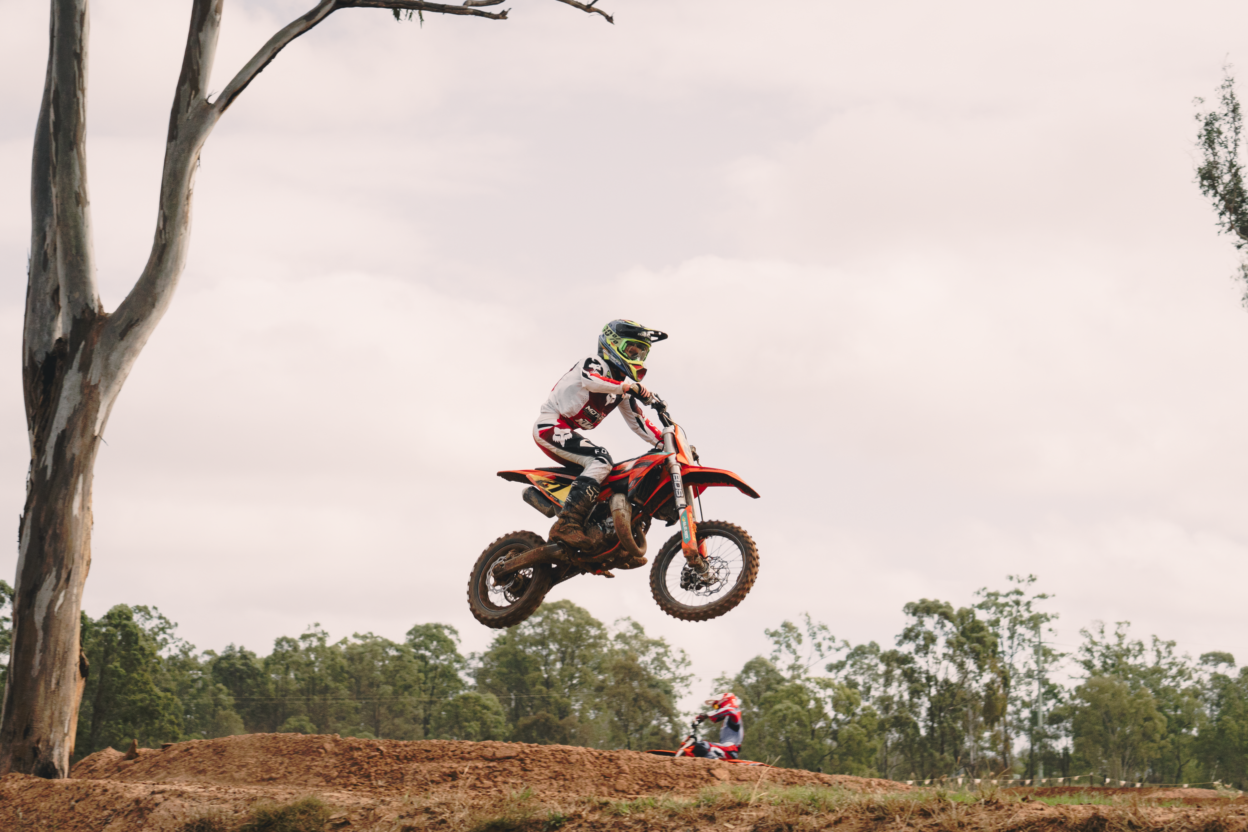 A motocross rider in racing gear riding an orange dirt bike in mid-air during a jump on a dirt track, with trees and cloudy sky in the background.