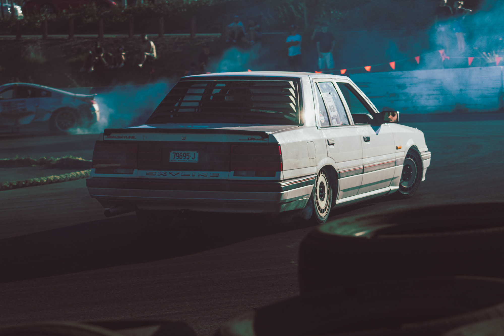 A white vintage car drifting around a corner on a race track with smoke behind the tires, while a second car is visible in the background. There are spectators watching from the side of the track.